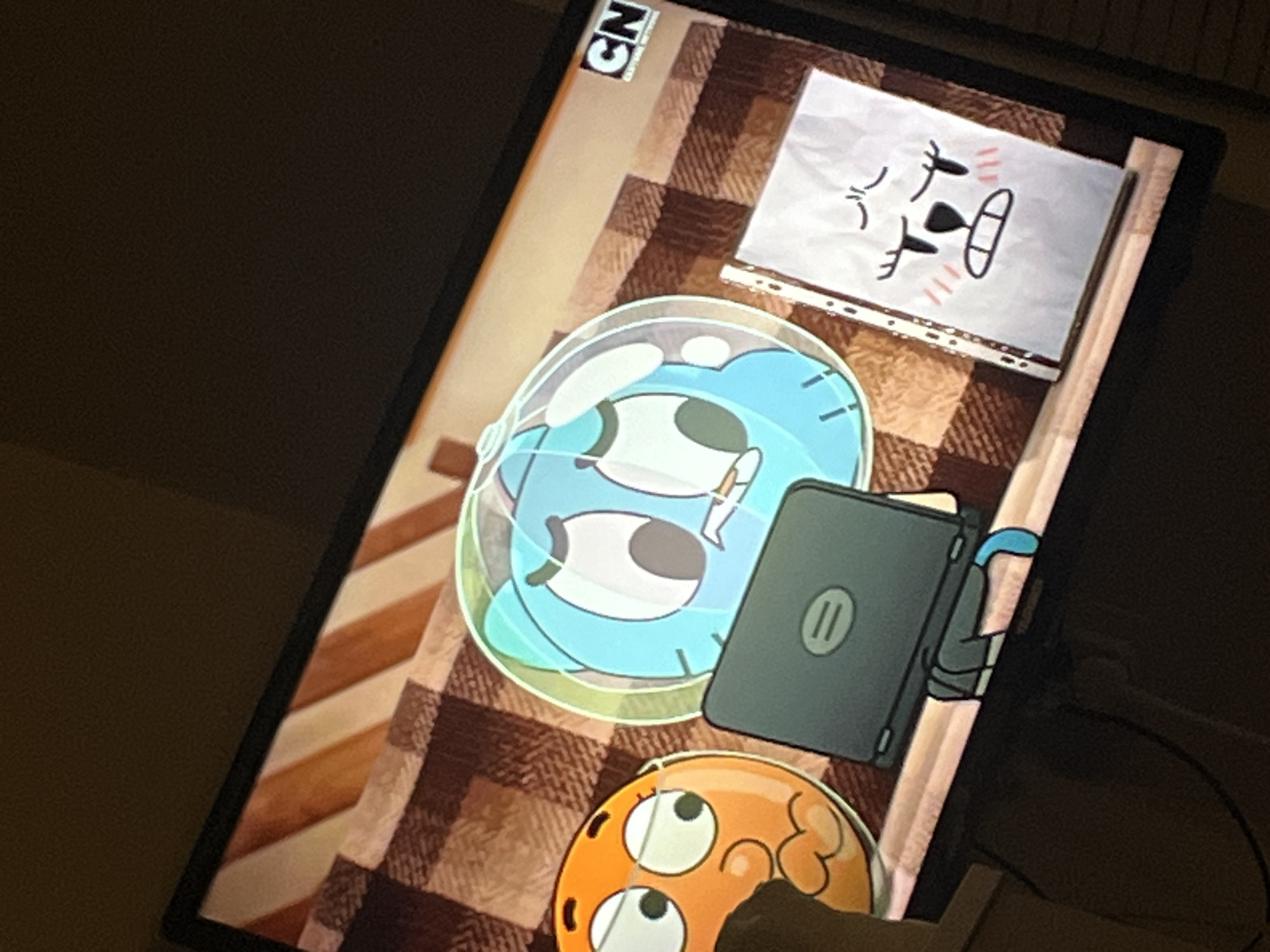 The image shows a television screen displaying an animated scene from "The Amazing World of Gumball." On the screen, two cartoon characters are visible: a blue cat-like creature with large eyes, enclosed in a clear bubble, and an orange, round creature with glasses, looking surprised. Between them is a laptop. To the right, a piece of crumpled paper with a drawing of a distressed face is propped up. The background of the animated scene is a patterned blanket. The television itself is set against a dark, out-of-focus background, likely a room. There is no information in the image to suggest its location in Biaga, Bulgaria.