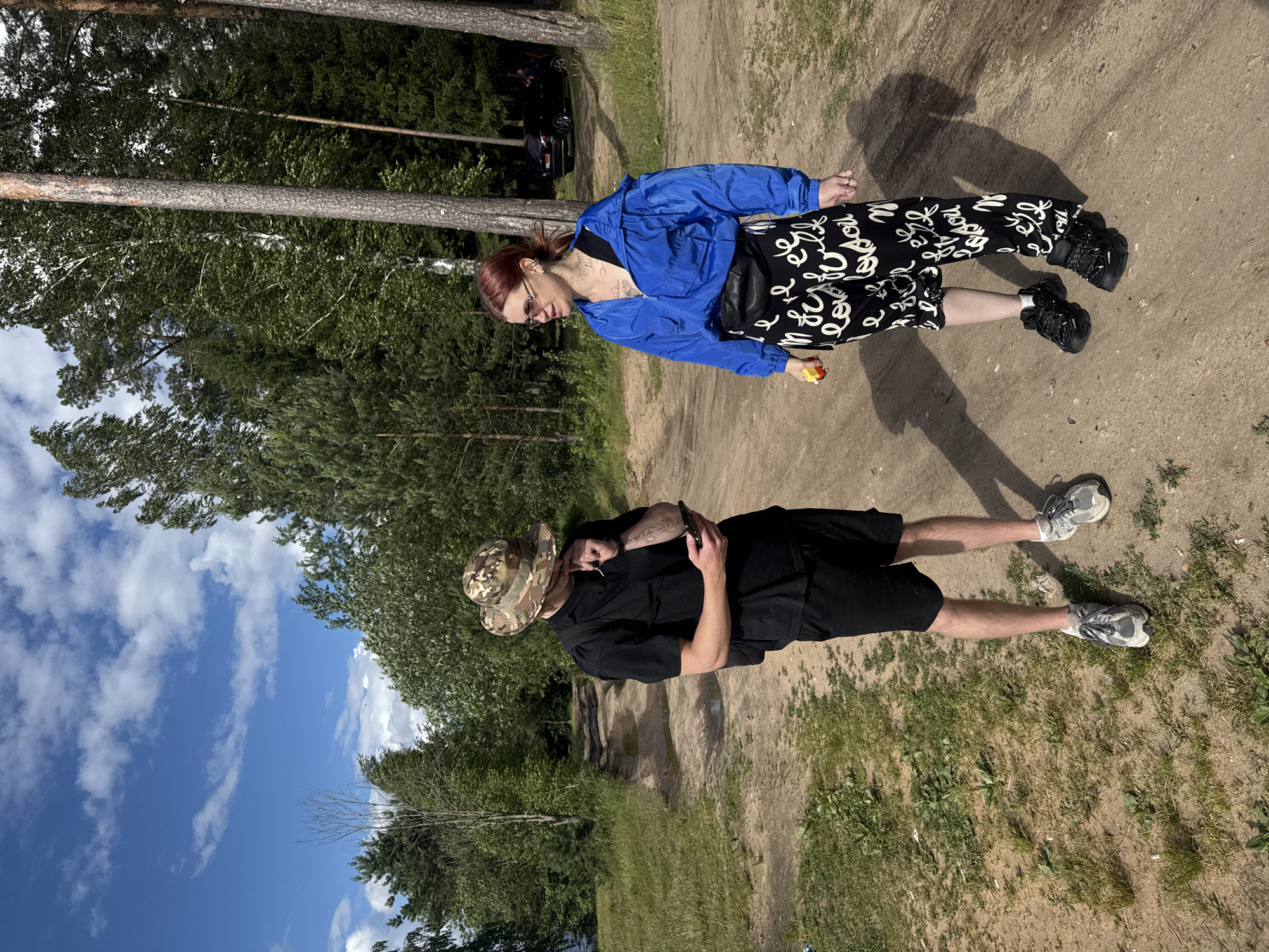 Two people are standing on a dirt path in a wooded area. The person on the left is a woman with red hair, wearing a blue jacket, black top, and black pants with white writing on them. She is wearing sunglasses and holding a small yellow object. The person on the right is a man wearing a camouflage hat, a black t-shirt, black shorts, and grey sneakers. He is holding a phone in his hand and has tattoos on his arm. The ground is a mix of dirt and grass, with trees and foliage in the background. The sky is blue with white clouds. The image appears to be taken on a sunny day, likely in the afternoon. Visible text includes writing on the woman's pants.