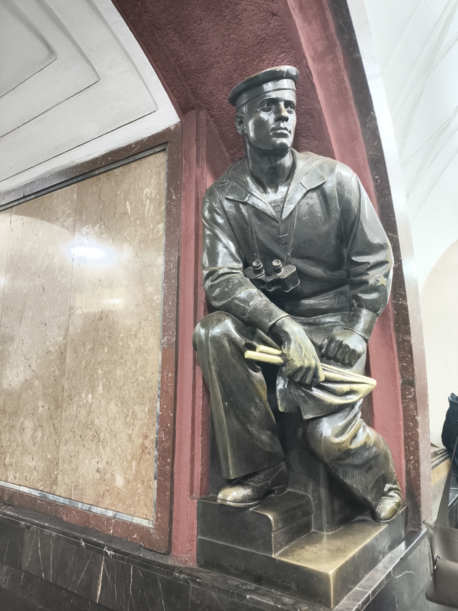 The central subject is a bronze statue of a male sailor, depicted seated and looking towards the right with a serious, contemplative expression. He wears a traditional sailor's uniform, including a peaked cap with "MAPT" visible in Cyrillic letters on the band, and a wide-collared shirt with binoculars hanging around his neck. His hands are clasped, holding a rolled-up item resembling a map or scroll, along with what could be a pair of pencils or small tools.
The statue is set within an architectural niche. Behind it is a dark red-painted wall. To the left, a large panel of polished light brown or beige marble with darker veins covers the wall. Below this marble panel and the statue's base is a dark, possibly black or very dark brown, marble wainscoting featuring prominent white veining. Above the marble panel, a white ceiling with layered, stepped molding is visible. The overall setting suggests an interior public space, potentially a grand metro station or a similar edifice.
The lighting appears to be artificial, creating bright reflections on the polished marble surfaces. The bronze statue has a subtle greenish patina in some areas, characteristic of aged bronze. The sailor's posture and gaze convey a sense of vigilance or readiness. The sculptural style and setting evoke Soviet-era artistic and architectural design.