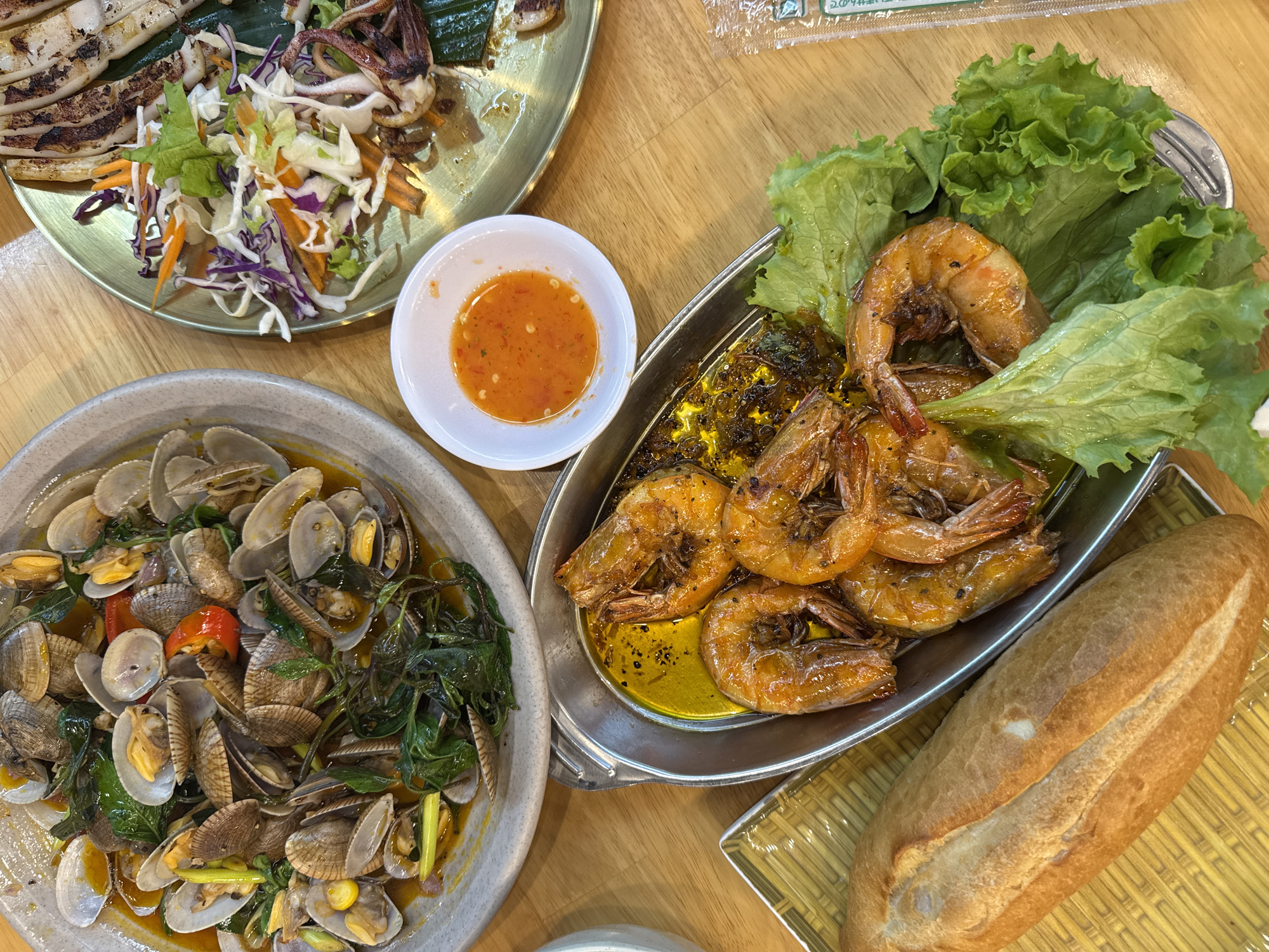 The scene depicts an overhead view of a wooden dining table laden with several dishes of prepared Vietnamese seafood, indicative of an eatery in Đà Nẵng, Vietnam.
In the upper-left, a rustic grey ceramic bowl contains numerous cooked clams in a reddish-orange broth, garnished with green leafy herbs and red chili slices. Many clams are open, revealing their interior.
To the upper-right, an oval, golden-hued metal platter displays strips of grilled squid with char marks, accompanied by a side salad of shredded purple cabbage, white cabbage, carrots, and green lettuce. Several grilled squid tentacles are integrated into the salad.
In the lower-middle, a silver-toned metal serving dish holds approximately seven large, grilled prawns submerged in a pool of yellow, seasoned oil with visible black specks, resting on a bed of fresh green lettuce leaves.
A small, white ceramic bowl positioned centrally contains a bright orange-red dipping sauce with visible chili flakes.
On the lower-left, a golden-brown baguette (bánh mì) is placed on a yellow, woven mat.
A clear plastic packet with Japanese text is partially visible on the far right edge of the wooden table. No individuals are present in the frame.
