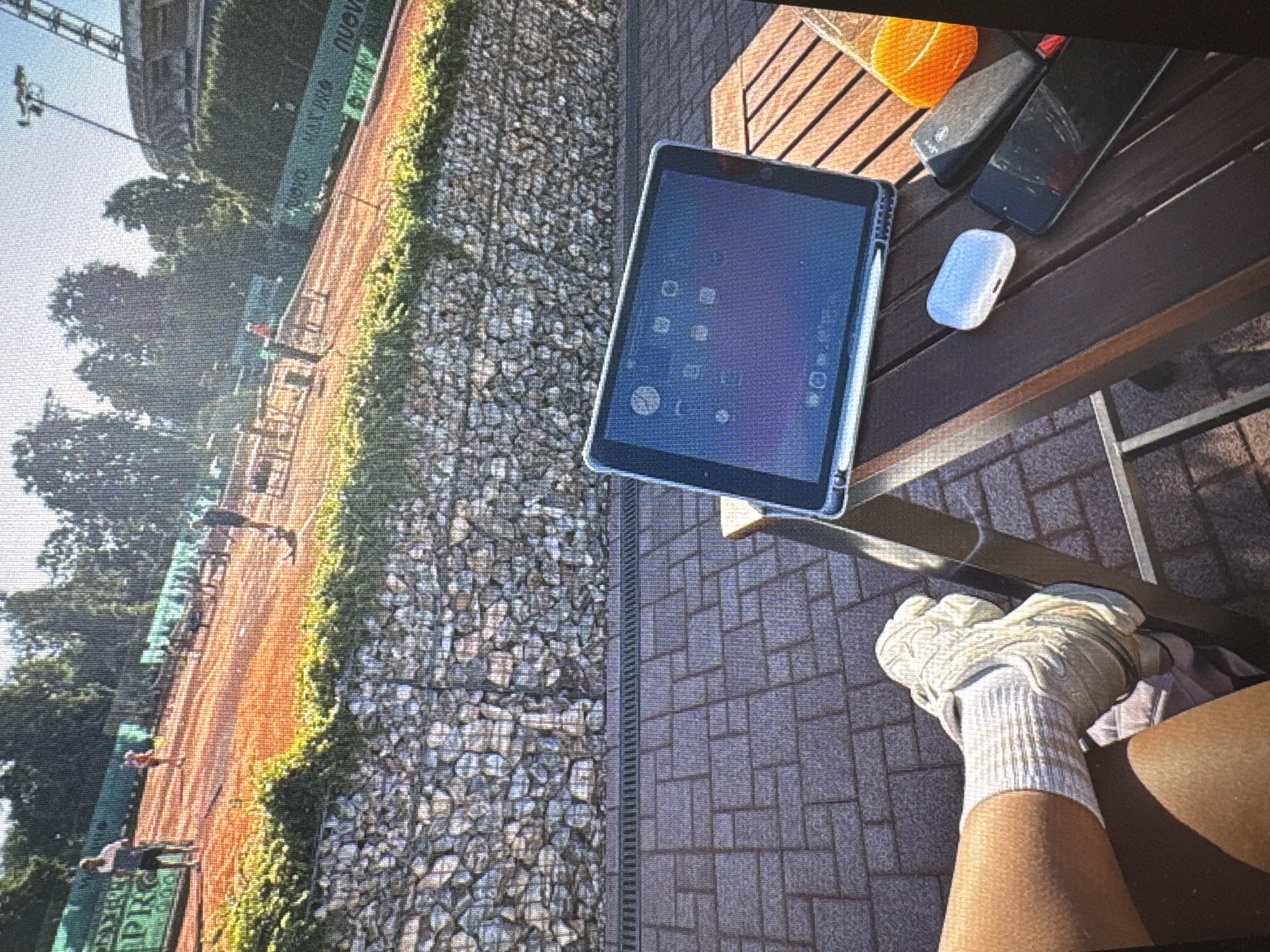 The image captures an outdoor tennis court scene in Vratsa, Bulgaria, viewed from an elevated perspective on a bright, sunny day. In the foreground, the lower arm and leg of a person are visible, featuring a white sock and a white tennis shoe or glove on the foot. This person is seated at a wooden table, on which various items are placed: a tablet with a stylus, a smartphone with a visibly cracked screen, a white wireless earbud case, an orange container likely for tennis balls, and a grey water bottle or thermos. The ground directly beneath the table is paved with dark grey interlocking bricks, separated from a gabion wall by a metal drain.
The gabion wall, filled with stones, runs horizontally across the midground and is partially covered with green foliage. Beyond this wall, a vibrant red clay tennis court is visible, where several individuals are actively engaged in playing tennis. At least four figures can be seen on the court, some mid-action. A green banner with white lettering lines the far side of the court, partially displaying the words "NUOVA" and "MAXIMA" (or "MAX MA"). Trees border the tennis court, and in the background, a large building or stadium structure is partially visible under a clear, bright sky. The strong sunlight casts distinct shadows, indicating daytime.