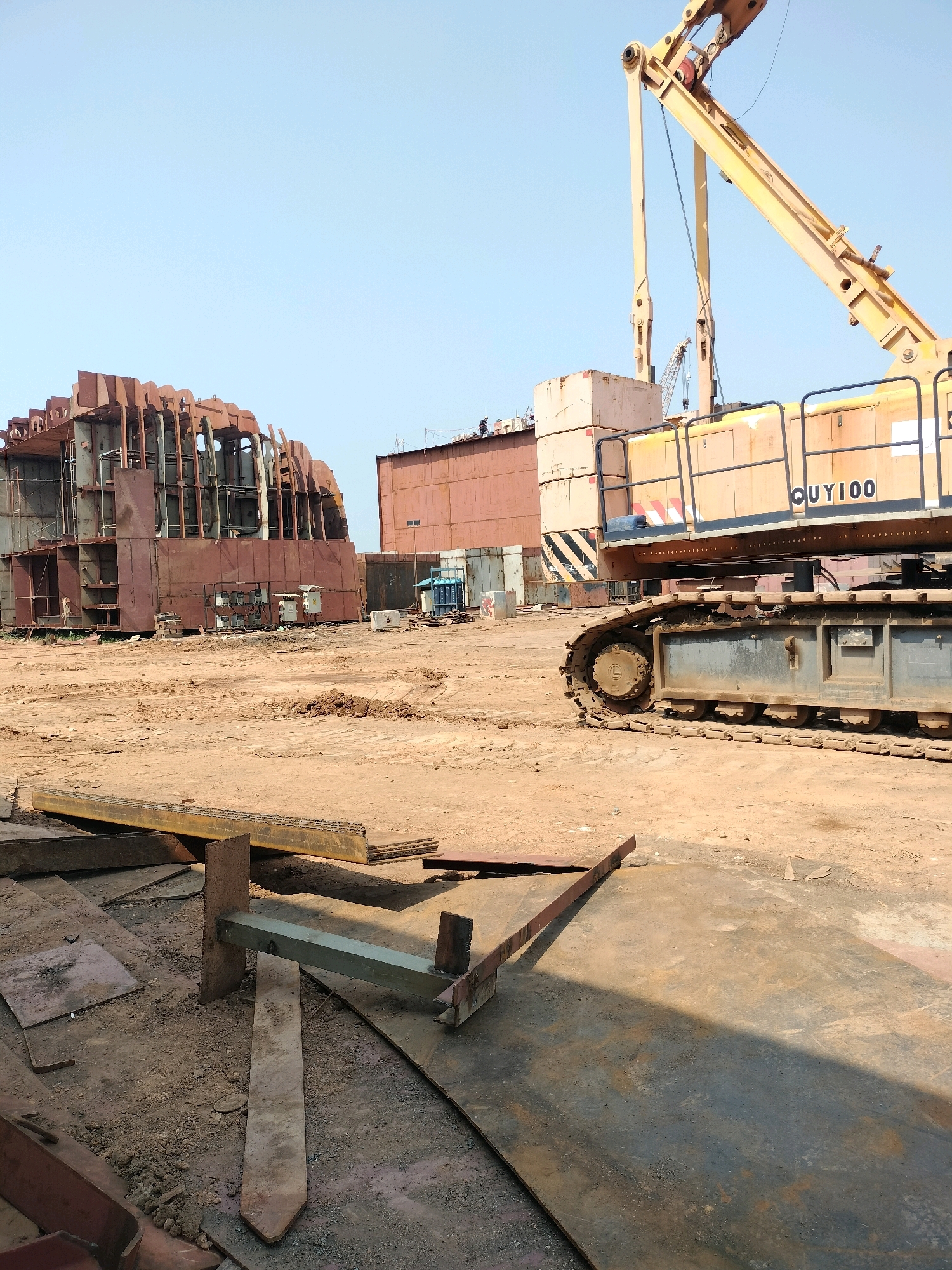 The image shows a shipyard in Batam, Indonesia, with several large, unfinished ship sections constructed of steel. A yellow crane with the designation "QUY100" is prominently featured, with its tracks and boom visible. The ground is composed of dry, sandy soil and large, flat pieces of metal. In the background, another ship hull segment and various containers are visible. Several construction workers can be seen on top of the ship structures, engaged in their tasks. The sky is clear and blue, indicating a sunny day. The scene depicts an active industrial area focused on shipbuilding.