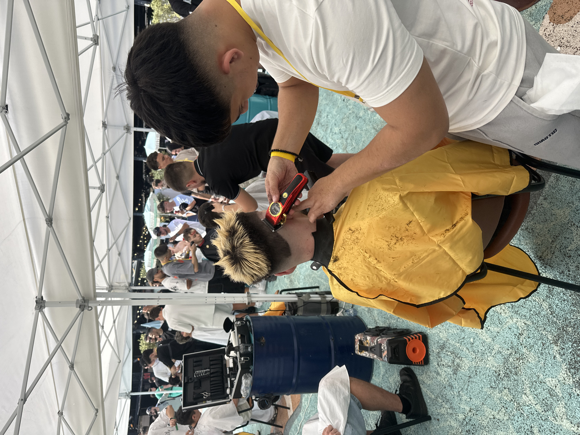 An outdoor scene, likely in Sofia, Bulgaria, depicts a barber event or competition under multiple white canopy tents during daylight hours. In the foreground, a male barber, wearing a white short-sleeved t-shirt with a yellow lanyard and grey sweatpants, is actively cutting the hair of a client seated with his back to the viewer. The barber holds a red and gold electric hair clipper in his right hand, focusing on the back of the client's head. A white tissue is tucked into the barber's right sweatpants pocket. The client wears a yellow barber's cape with black trim, and his hair features bleached blonde spikes on top with a dark fade on the sides and back.
To the left of the central figures, another individual, partially visible and wearing a white barber's gown, sits beside a blue metal barrel. The barrel serves as a stand for an open black toolbox-style case containing various barber tools, including scissors, combs, and spray bottles. A black and orange portable speaker is visible on the light blue speckled ground near the client's feet.
In the mid-ground and background, numerous individuals, primarily men, are engaged in similar barbering activities or observing. Several barbers are visible under additional white tents, some wearing white gowns and actively cutting hair. The ground surface is a light blue, textured material with white speckles. The distant background reveals more white tents, light blue market umbrellas, and green foliage under a clear sky.