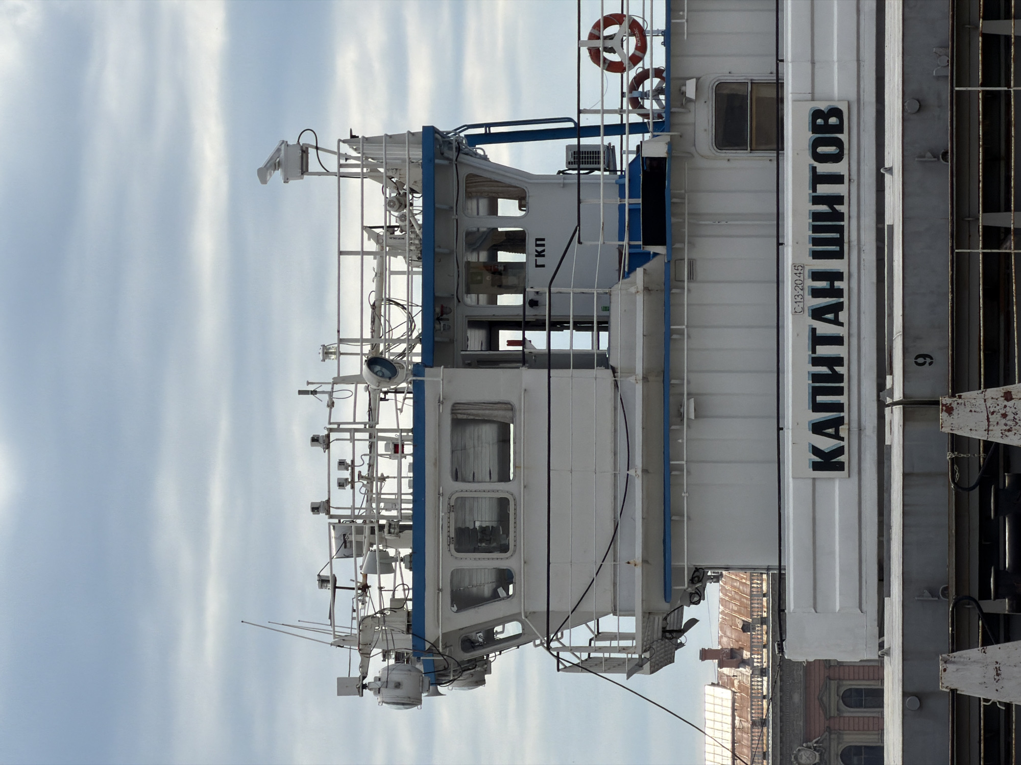 The image, when viewed in its correct orientation (rotated 90 degrees counter-clockwise), displays the side view of a large white vessel, likely a ship or ferry, against a light, cloudy sky. The vessel's superstructure is predominantly white with blue railings and accents.
The main subject is the middle to upper deck section of the vessel, showcasing its bridge or wheelhouse area. This section features multiple rectangular windows, some with reflections, and an air conditioning unit is visible on an upper deck. The vessel is equipped with various maritime instruments and apparatus, including numerous antennas, radar units, searchlights, and other communication devices mounted on its mast and upper deck structures. Two orange and white life rings are visible on the stern side of the superstructure.
Prominent Cyrillic text is visible on the vessel's side. The largest lettering spells out "КАПИТАН ШИТОВ" (KAPITAN SHITOV), indicating the vessel's name. Below this, a smaller inscription reads "C 13-20-45". Another abbreviation, "ГКП" (GKP), is displayed on the wheelhouse section.
The setting appears to be daytime, with a bright sky characterized by soft, elongated cloud streaks, suggesting either an overcast day or diffused sunlight. In the distant background, to the right of the vessel, parts of multi-story buildings with classic European architecture, including arched windows and reddish-brown roofs, are discernible, indicating the vessel is likely docked or near a city waterfront. The overall scene suggests a calm, unhurried atmosphere, with no immediate activity or people visible on board. Given the location context, these buildings are likely in Saint Petersburg, Russia.