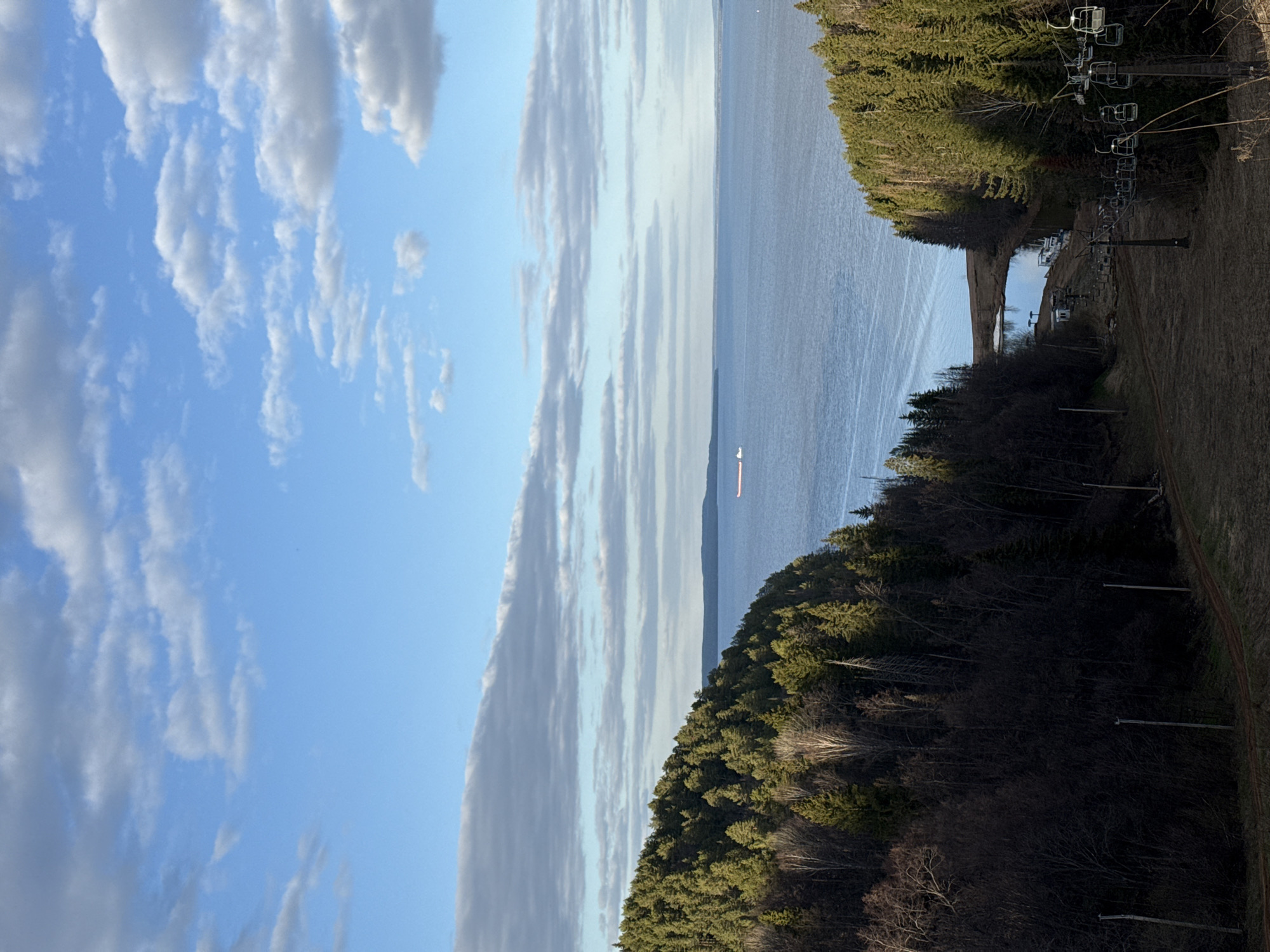 The image depicts a landscape from an elevated position, possibly a hill or mountain, overlooking a body of water. The sky is partly cloudy, with a mix of blue sky and white clouds. On either side of the water, there are forested areas with green trees. On the right side of the frame, there is a ski lift structure with chairs. The terrain on the right side appears to be a ski slope. A ship can be seen sailing at the horizon in the body of water. The image is taken in Galevo, Russia.