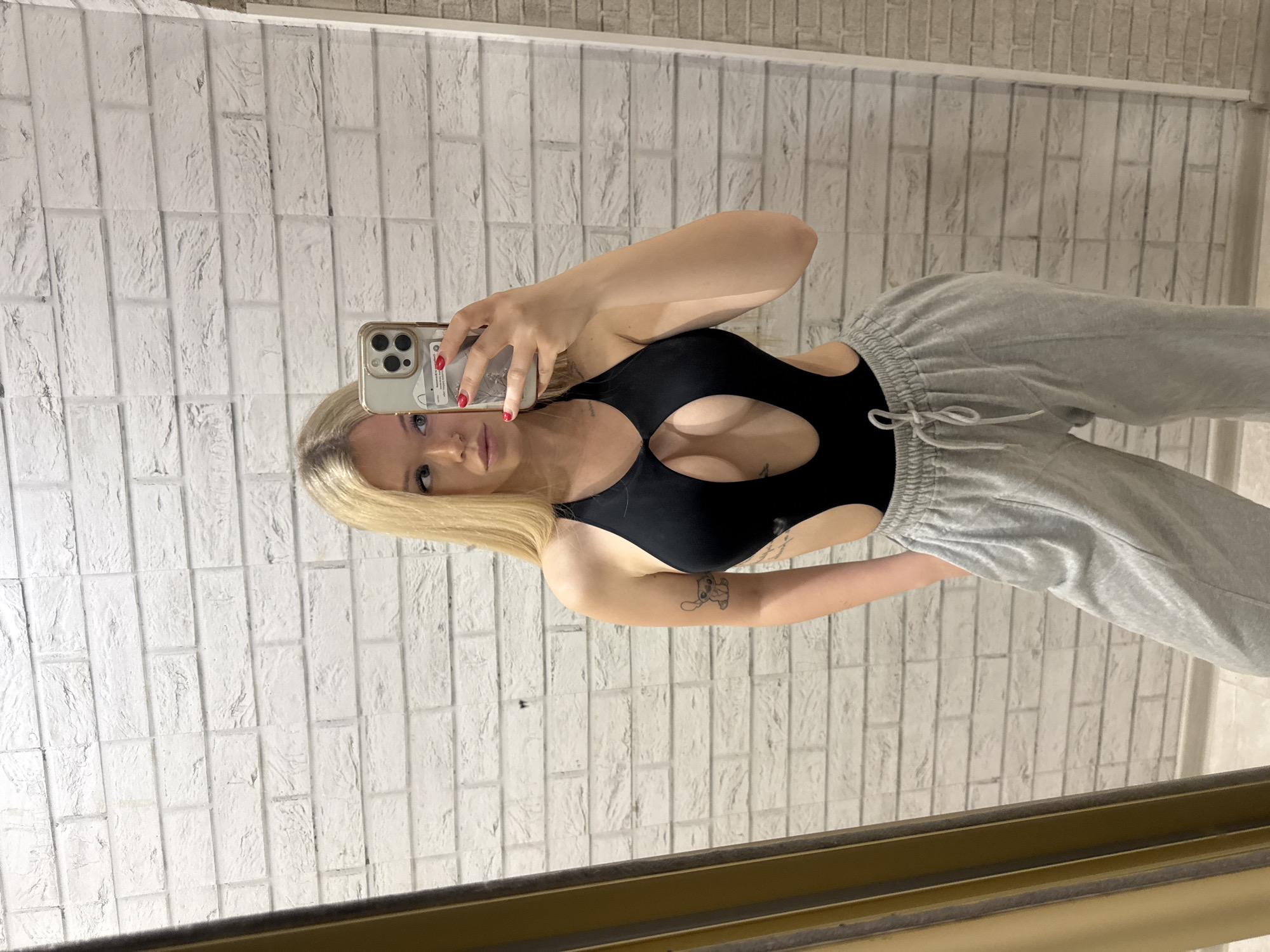 A young woman with blonde hair is taking a mirror selfie in what appears to be a residential setting. She is wearing a black one-piece swimsuit with cutouts on the chest and sides, and grey sweatpants. She is holding a smartphone in her right hand and is looking at her reflection in the mirror. The background is a white brick wall. The image does not contain any elements that would allow for the determination of the location in the city of Saint Petersburg, Russia, or any other specific city.