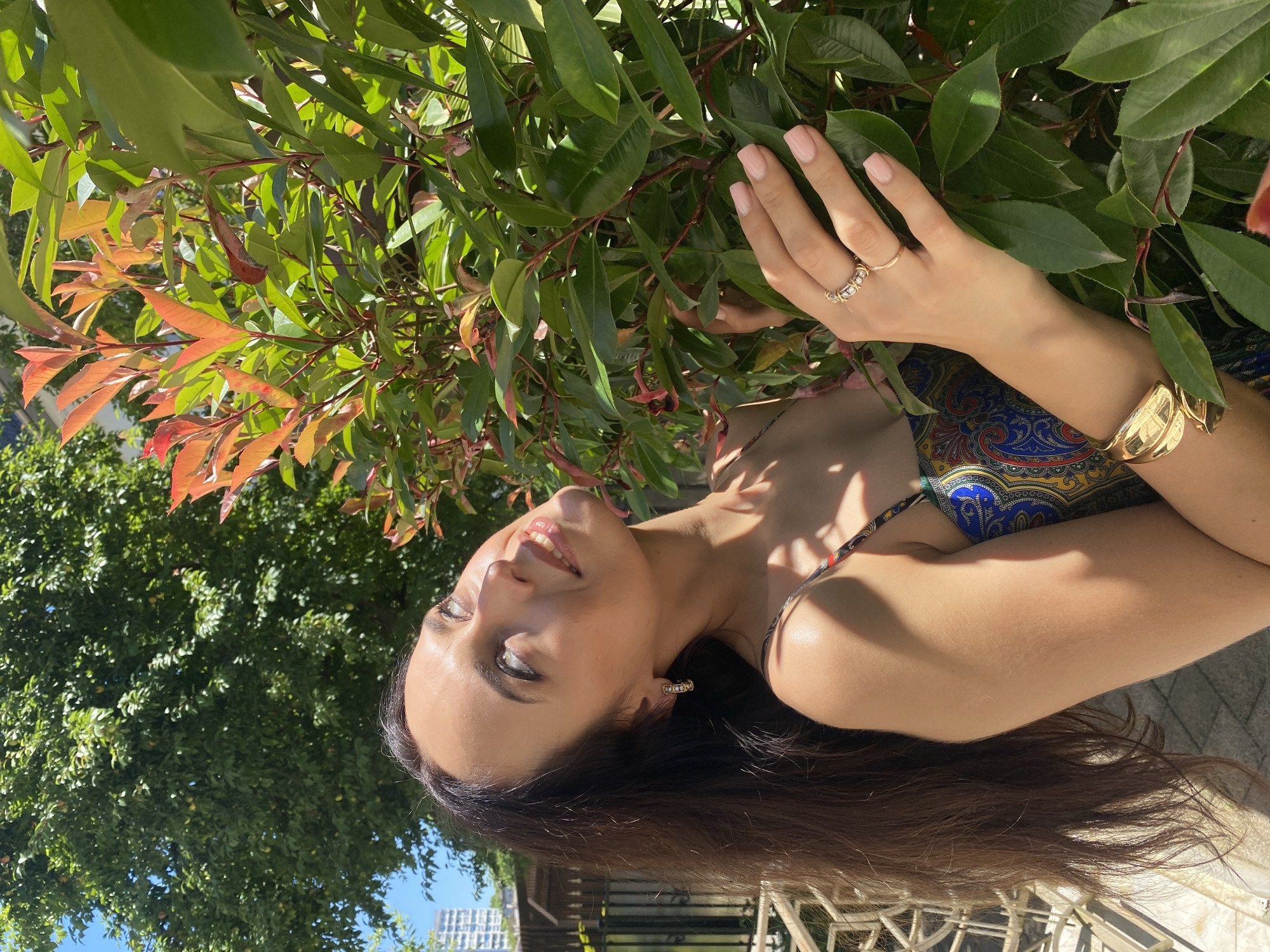 A woman with long, dark hair is looking upwards towards a vibrant green and reddish-orange plant. She is wearing a patterned dress, a gold bracelet, and rings on her right hand. The scene appears to be outdoors, with sunlight dappling through the leaves and casting shadows on her arm. In the background, a hint of a city skyline and a fence are visible, suggesting an urban setting. It is plausible this photograph was taken in Sochi, Russia, given the vegetation and architectural elements often found in the city.