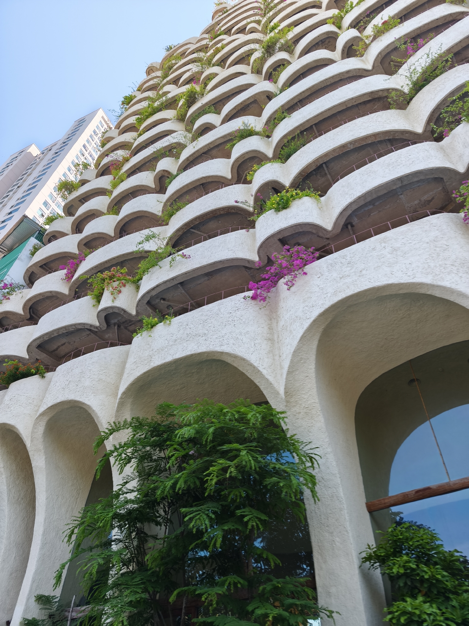An exterior, low-angle shot showcases the unique architectural design of a building in Nha Trang Ward, Vietnam. The building features a series of staggered, crescent-shaped balconies, each adorned with lush green plants and vibrant pink bougainvillea flowers. The balconies are made of a rough, textured white material, creating a distinctive pattern as they recede upwards. Below the balconies, the building has large, arched entrances. Green trees and foliage are visible at the base of the building, partially obscuring a large glass window. To the left, a taller, more modern white building with many windows stands in contrast to the organic design of the main structure. The sky is a clear, pale blue.