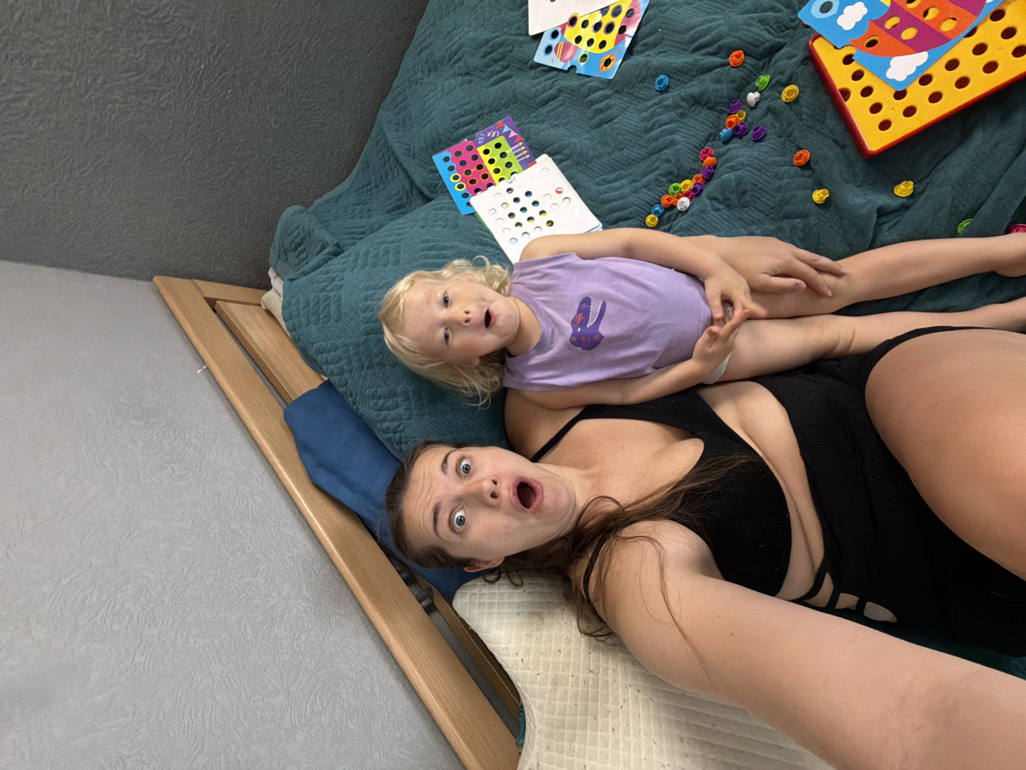 A woman and child are lying on a bed. The child is fair-haired and wearing a light purple shirt with a reptile design on the front. The woman is wearing a black outfit. Both the woman and the child have their mouths open in a surprised expression. The bed has a green quilt with several children’s toys scattered on it. The bed is on a wood frame, and the floor is grey. Based on other information sources, the location is likely to be Sochi, Russia.