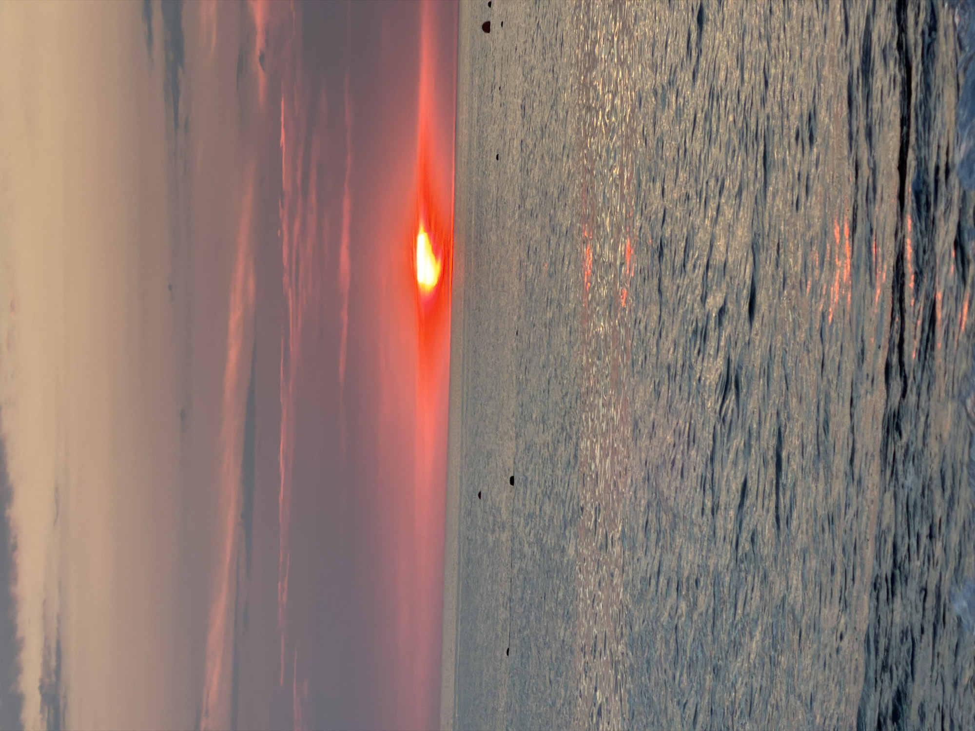 A vertical, full shot captures a sunset over the Black Sea in Sochi, Russia. The sky is a gradient of soft pink and grey clouds, transitioning to a vibrant orange and red near the horizon where the sun is setting. The sun itself is a bright, fiery orb peeking just above the water. The sea is calm with gentle waves, reflecting the colors of the sunset. Several dark, spherical buoys are visible in the water, scattered across the surface. The foreground shows the edge of the water with small waves breaking.