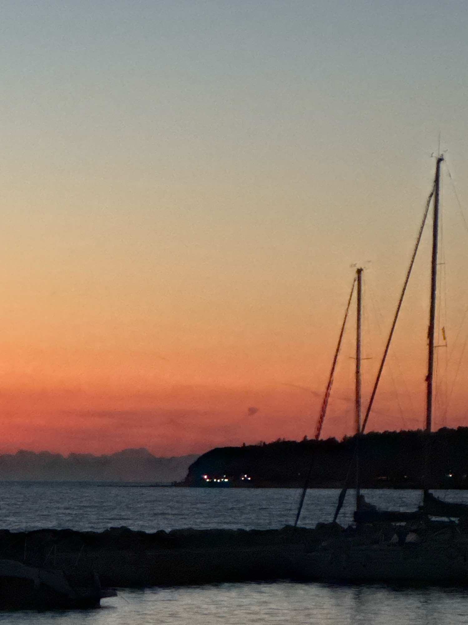 The image shows a twilight scene with a gradient sky transitioning from pale blue at the top to a vibrant orange and red near the horizon. The calm sea reflects the sky's colors. In the foreground, dark silhouetted objects likely represent a breakwater or shore made of rocks, with a few indistinct shapes of boats moored in the water. In the middle ground, a dark, tree-covered coastline juts into the sea, with a few small lights visible on its edge, suggesting a settlement. Dominating the right side of the image are the dark silhouettes of two sailboat masts, one taller than the other, reaching towards the sky. The overall impression is one of quiet evening by the sea, with the natural beauty of the sunset contrasting with the man-made structures of the boats and distant lights.