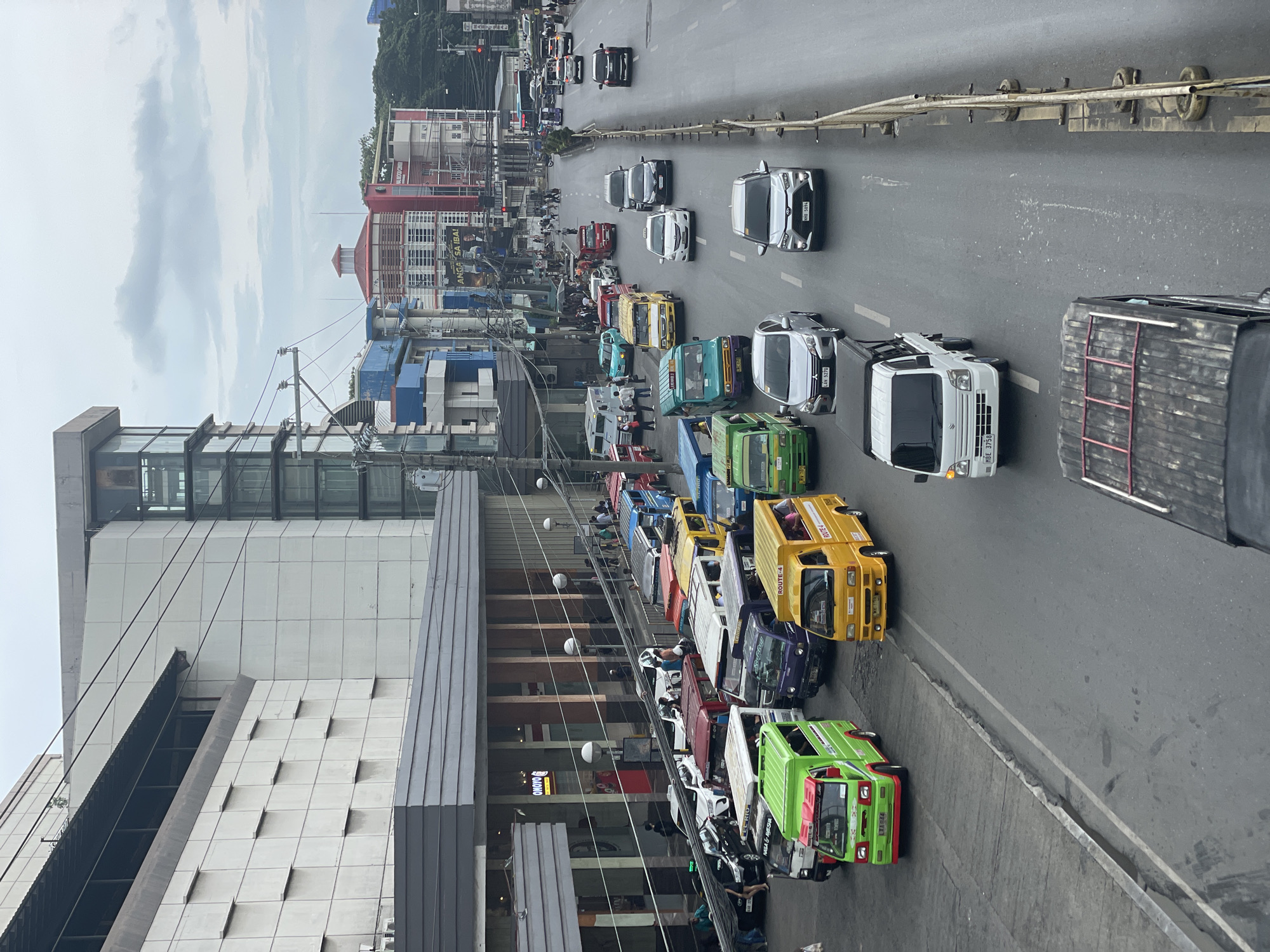 The image depicts a street scene in Davao City, Philippines, likely near a commercial area, with a multi-level building on the left and a busy road filled with various vehicles. The building on the left features a modern design with a textured facade and large windows. Across the street, a mix of colorful public utility vehicles, including jeepneys and vans, are seen navigating the road. Several cars and trucks are also present, indicating moderate traffic flow. Pedestrians are visible on the sidewalk near the building and crossing the street, suggesting a lively urban environment. The sky is overcast, hinting at a cloudy or possibly rainy day. The overall scene captures the dynamic transportation and commercial activity characteristic of a Philippine city.