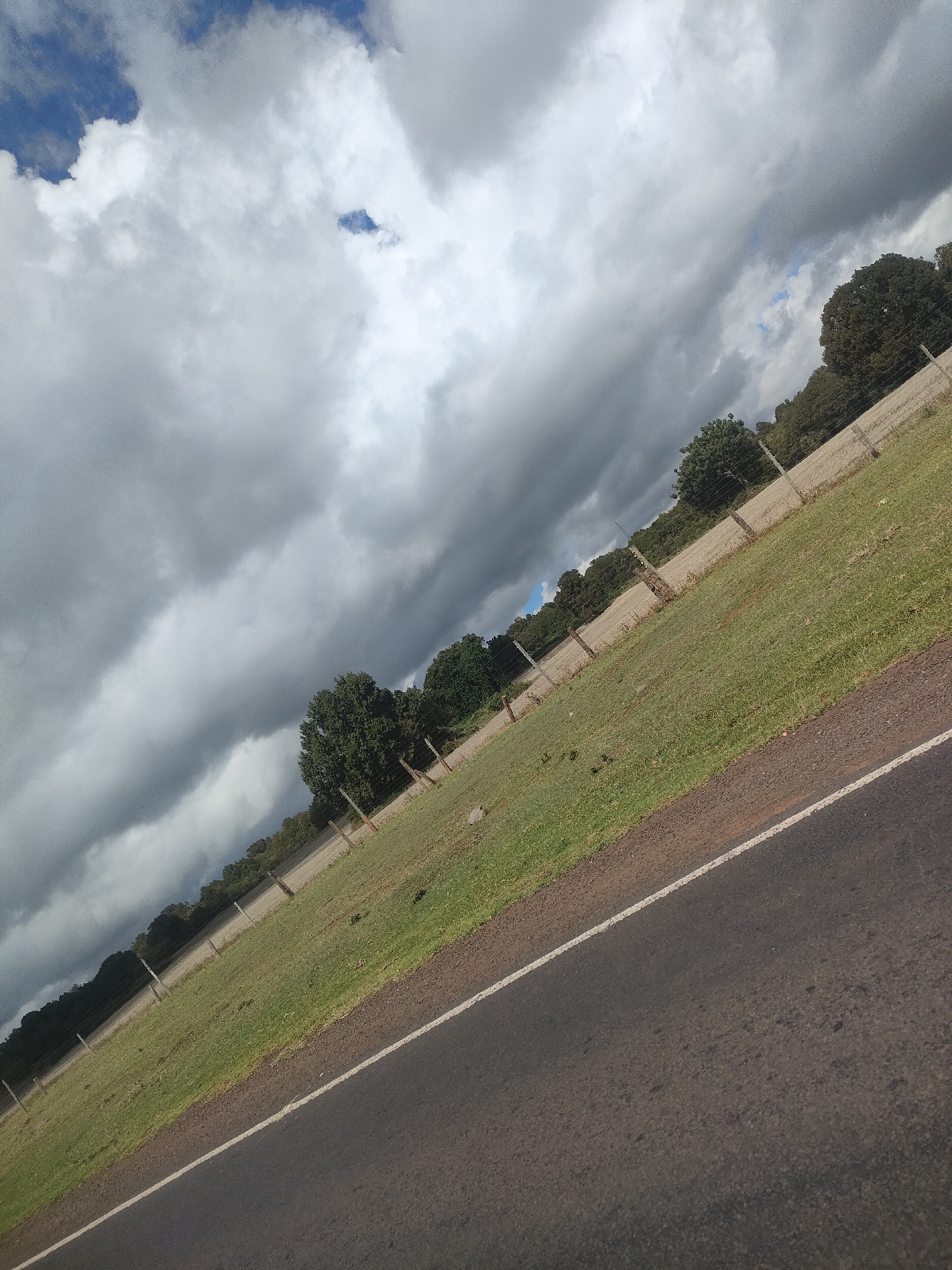 The image depicts a road in a rural area. The road is made of asphalt and has a white line marking the edge. To the left of the road is a grassy field with trees and a fence along the edge. The sky is filled with dark, ominous clouds, suggesting an approaching storm. There are no people or other subjects visible in the image. The location is Kisima ward, Kenya.
