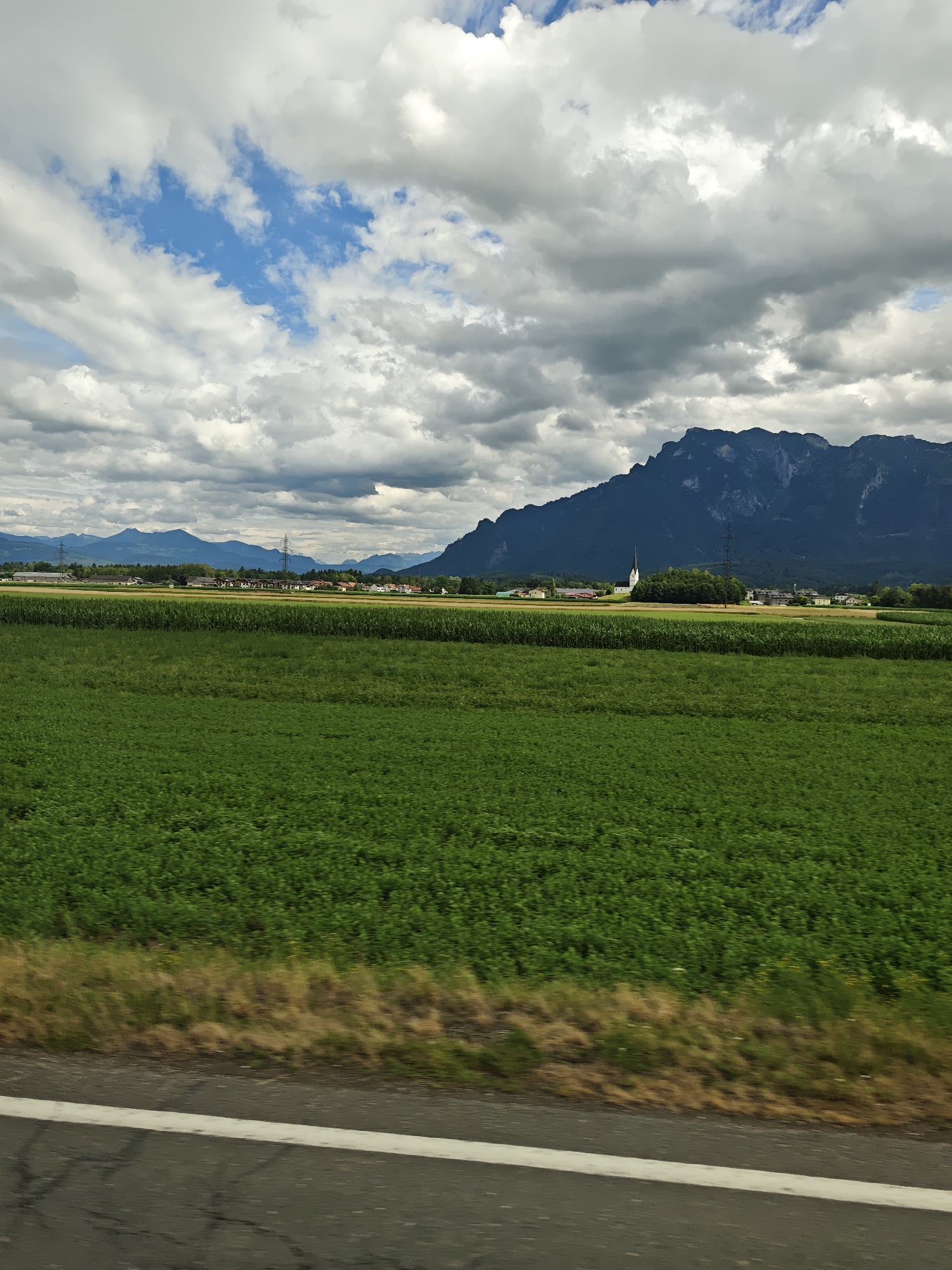 The image captures a panoramic view of a rural landscape under a cloudy sky, likely taken from a moving vehicle on a road. The foreground is dominated by a dark asphalt road with a white line, suggesting motion blur. Adjacent to the road is a strip of dry, brown grass. Beyond this, a vast expanse of vibrant green fields stretches out, possibly crops or pastures. In the mid-ground, a line of trees partially obscures a collection of buildings, including a white church with a steeple, indicative of a village or small town. Further in the distance, more fields and trees are visible, leading towards a range of imposing, dark mountains that form the backdrop of the scene. The sky is a dramatic mix of bright blue patches and heavy, grey clouds, hinting at changeable weather. The overall impression is one of a tranquil, agricultural area with a majestic mountain range.