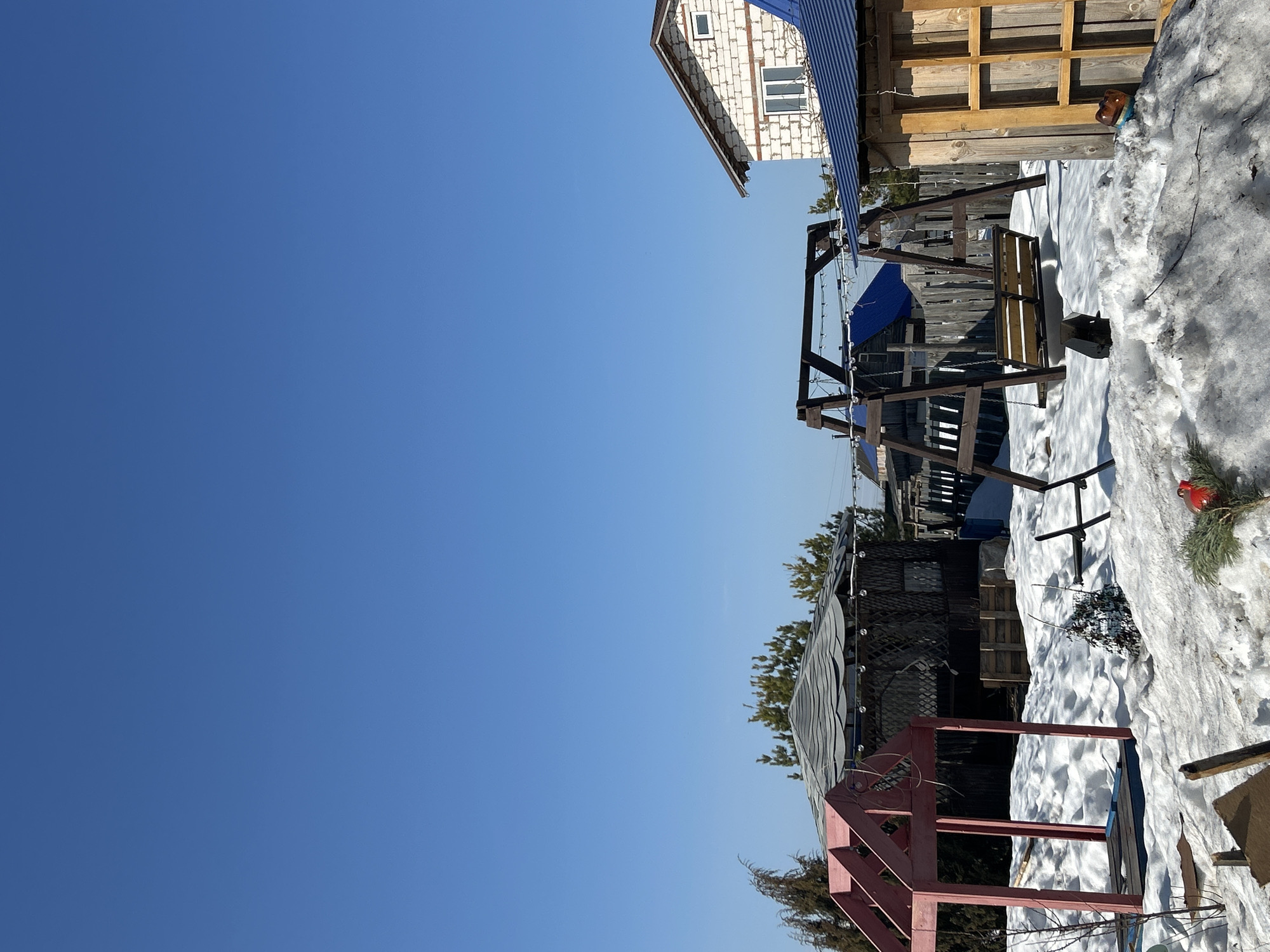 The image depicts a winter scene, likely in the vicinity of Ольховка, Russia, given the presence of buildings and structures typical of the region. A clear blue sky dominates the upper half of the image, contrasted by the lower half containing snow-covered ground and structures.
To the right of the image frame, the edge of a multi-story building with what appears to be white brick or siding and visible windows is apparent. Adjacent to this building is a wooden structure, likely an outbuilding or shed, with exposed timbers and stacked wooden panels. Snow has accumulated around the base of these structures.
A swing set stands prominently in the middle of the frame. The swing set appears to be constructed from dark-stained wood with a bench-style swing hanging beneath. Additional wooden structures are situated behind the swing set, potentially fencing or other outbuildings, partially obscured by the swing set frame.
Near the bottom of the image, a small red-painted structure of indeterminate purpose can be seen. Snow is present around the base of this structure as well. The presence of a pine tree suggests the area is residential.