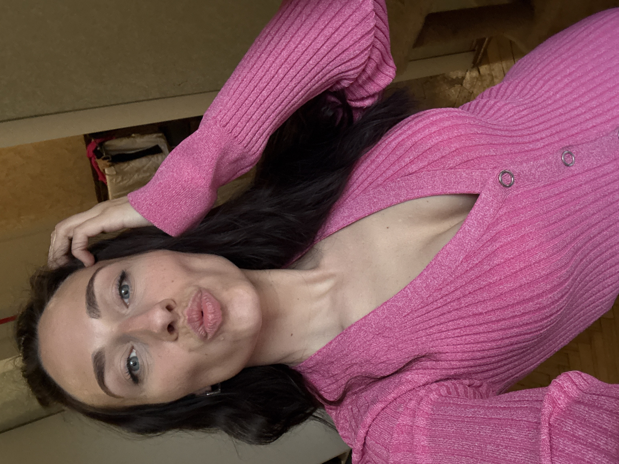 A woman with light eyes and dark hair is posing for a selfie indoors. She is wearing a pink ribbed top with metallic ring details. Her lips are pursed in a kiss. The background is blurred and shows a glimpse of furniture. The location cannot be determined from the image.