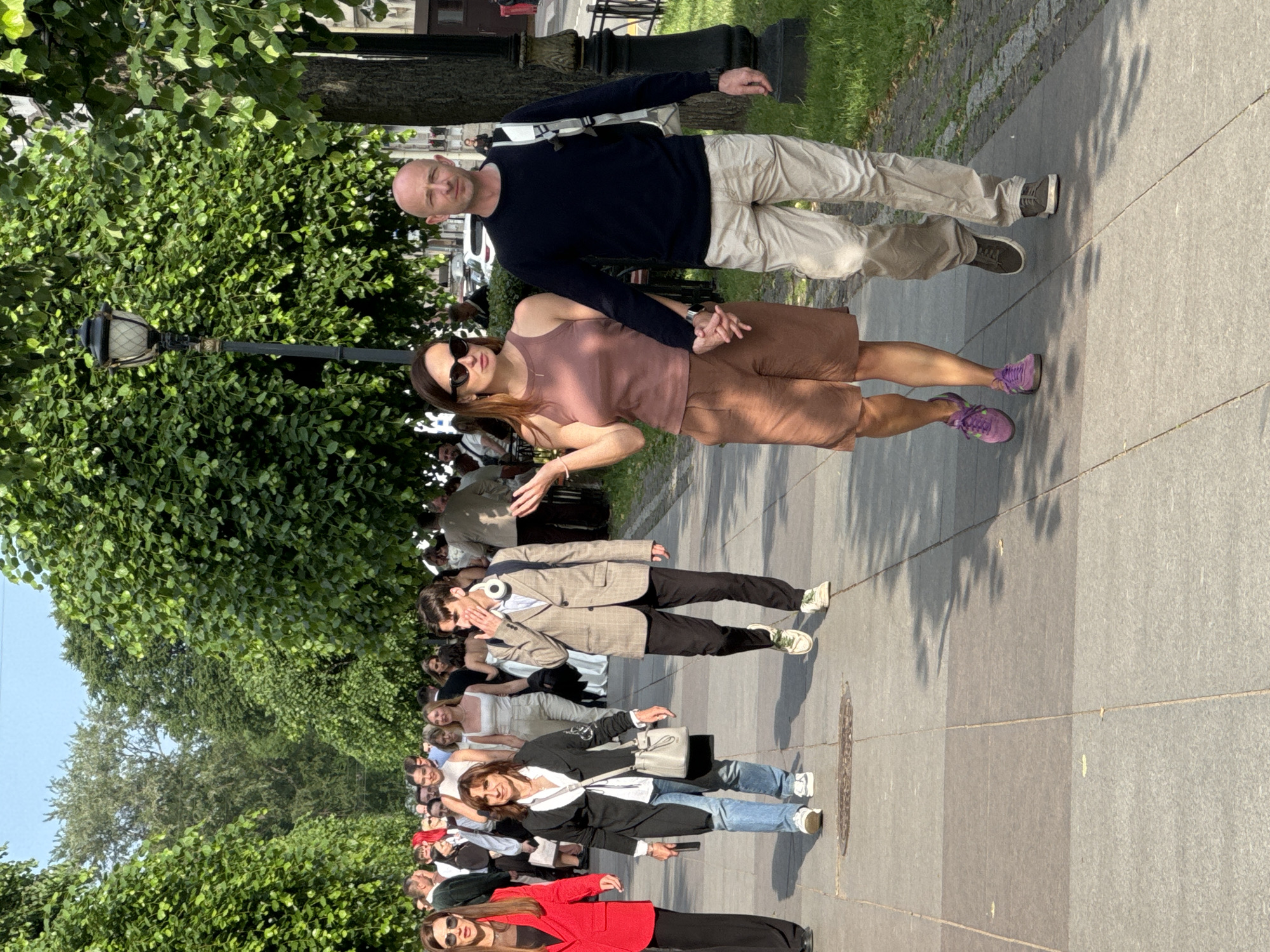 A group of people are walking on a paved walkway lined with trees. In the foreground, a man and a woman are walking hand-in-hand. The woman is wearing a sleeveless brown top and brown shorts, with purple sneakers. The man is wearing a navy blue long-sleeved shirt and light-colored pants, with dark sneakers. Behind them, other people are walking in the same direction. The walkway is made of stone pavers and there is a grassy area with trees to the right. The background shows more trees and buildings. This scene appears to be in Saint Petersburg, Russia, likely along a park or promenade.