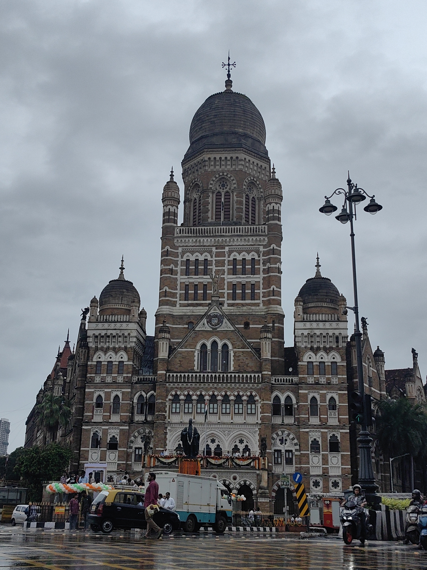 The image depicts a frontal view of the Mumbai City Hall (also known as BMC Building or Brihanmumbai Municipal Corporation Building) in Mumbai, India, under a cloudy sky. The grand Victorian Gothic Revival architecture of the building is prominent, featuring intricate detailing, multiple towers, and a large central dome. In the foreground, the wet street, likely from rain, reflects the surrounding environment and the traffic. Several vehicles, including a black and yellow taxi and a white generator van, are present on the road. A few individuals are visible near the vehicles and the building's entrance, with some appearing to be engaged in conversation or movement. A traffic light pole with multiple lamps stands to the right of the building. The scene captures a street-level perspective of this iconic Mumbai landmark on a possibly overcast day.