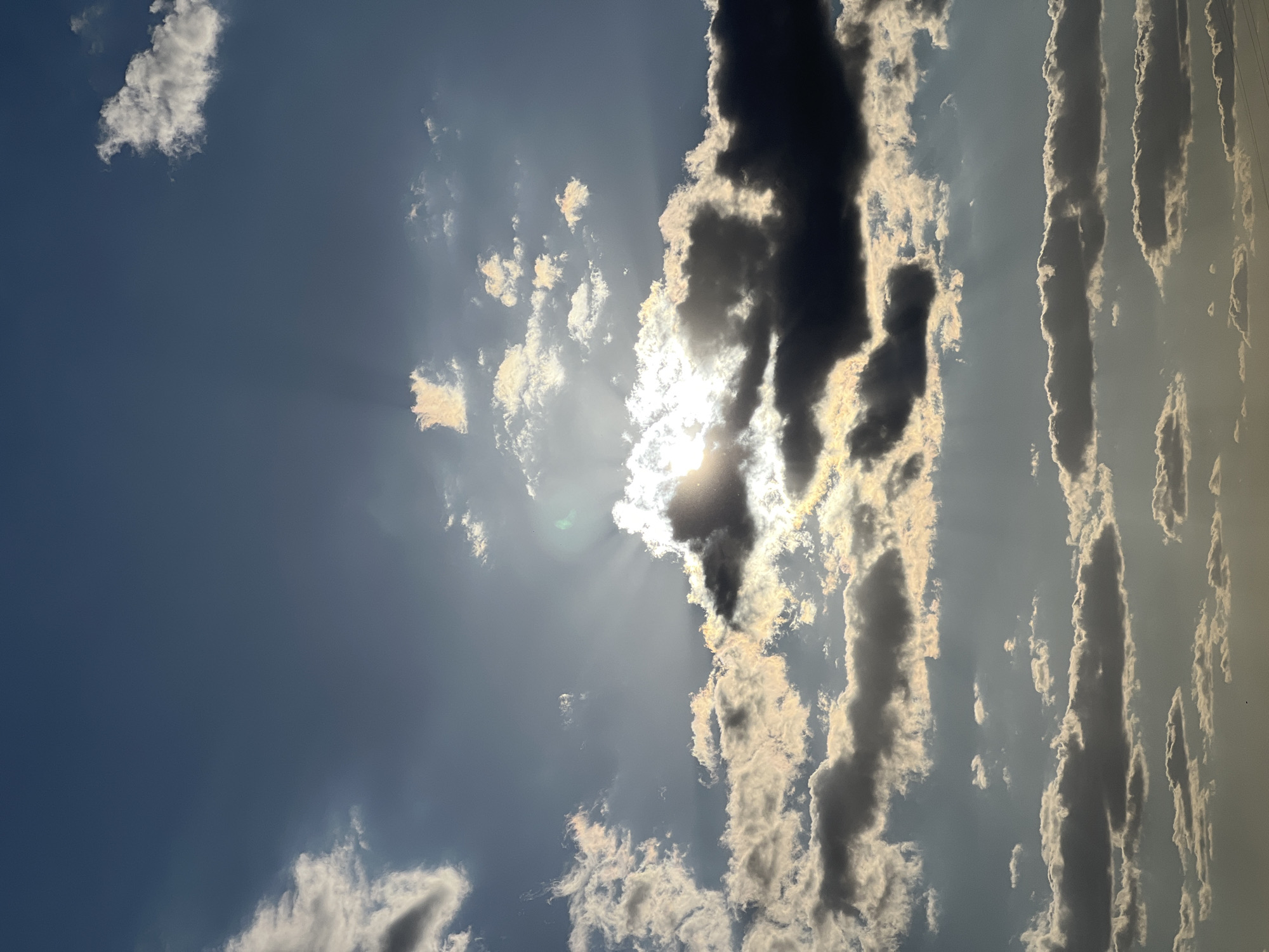 This is a vertical, close-up, eye-level shot of a partly cloudy sky. The sun is bright and visible, partially obscured by dark, thick clouds that are backlit by the sun's rays, creating a dramatic effect. There are scattered lighter clouds higher up in the frame. The overall color palette is blues, grays, and whites, with hints of yellow from the sun. The image does not contain any recognizable landmarks or details that would indicate a specific location within the city of Žatčany, Czechia. No people, subjects, objects, or actions are visible in the frame.