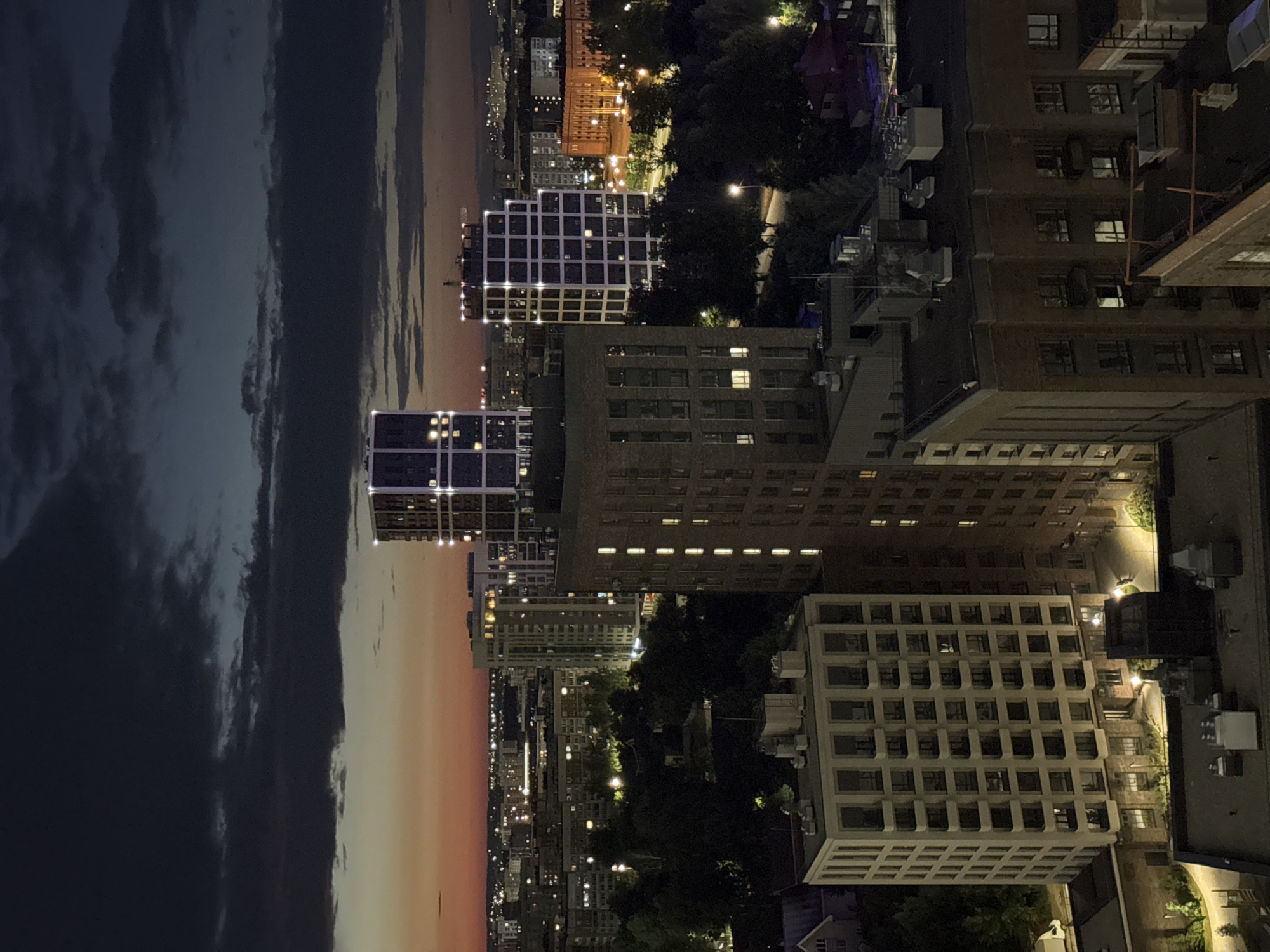 This is a night aerial view of a city skyline in Izhevsk, Russia, likely taken during dusk or early evening. The sky is a deep blue and black with dark, dramatic clouds, and the horizon is illuminated by a faint orange and pink glow from the setting sun or city lights.
The scene is dominated by various buildings, primarily multi-story residential complexes and modern architectural structures. In the foreground, a light-colored, boxy building with many windows and balconies is visible on the left. To its right, a taller, darker brick-colored building stands, also featuring numerous windows, some of which are illuminated. Further into the background, several more modern, illuminated high-rise buildings stand out. One particularly striking building has a distinctive white grid-like facade with bright lights emanating from within. Other buildings in the distance are illuminated with warm, yellow lights.
The urban landscape is interspersed with dark masses of trees, indicating the presence of parks or green spaces within the city. Streetlights are visible along roads and pathways, casting pools of light. The overall impression is one of a modern, developing urban environment at night, with a mix of older and newer architectural styles. There are no discernible people or specific actions captured in this high-level, distant view. The image focuses on the architecture and the atmospheric lighting conditions of the city.