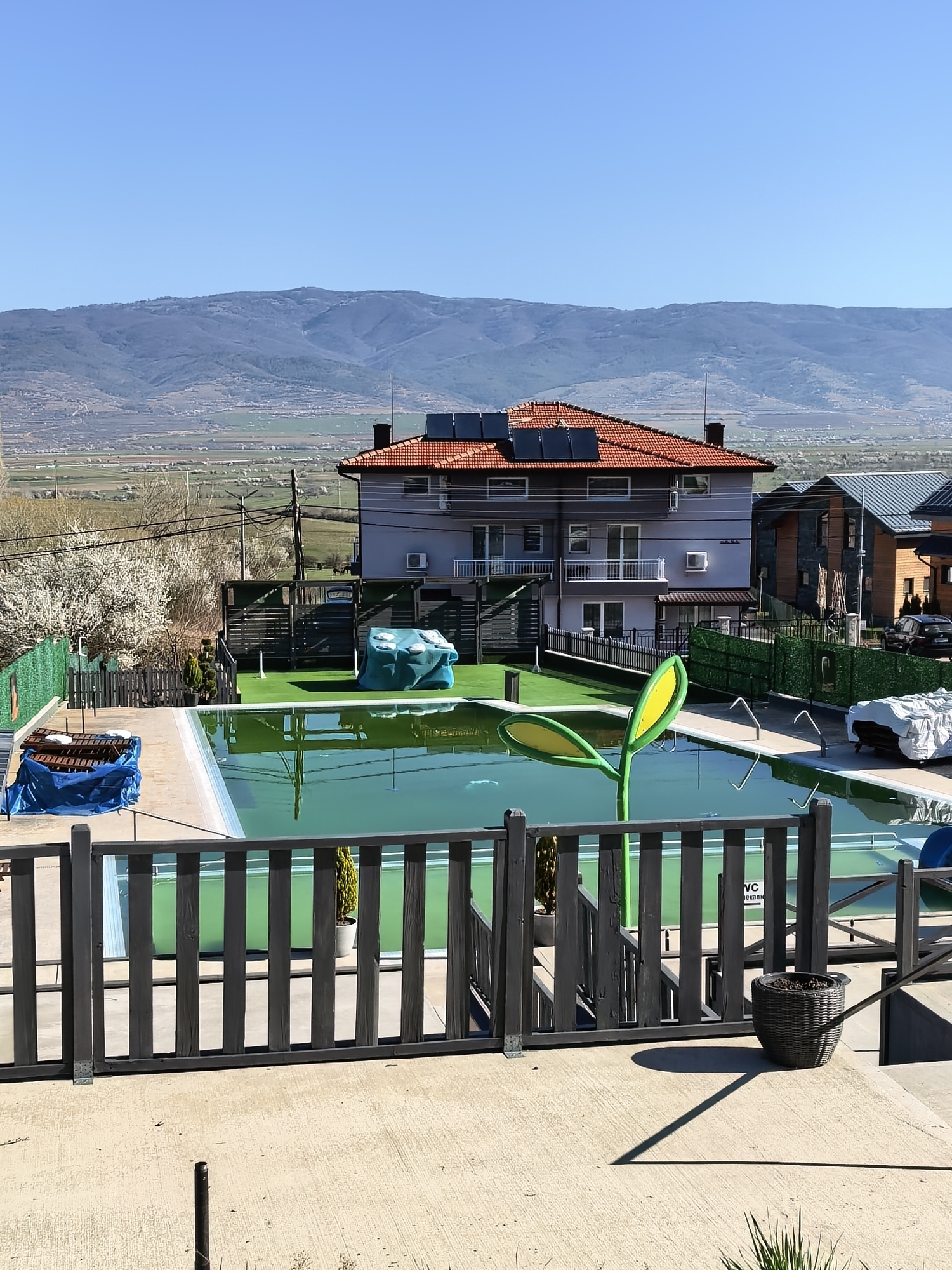 The image depicts an outdoor setting in a seemingly residential area in the city of Unknown, Unknown, with a swimming pool in the foreground. The pool water appears green, potentially indicating algae or water treatment issues. A wooden fence encloses the pool area, with a concrete patio extending beyond the fence.
In the middle ground, there's a lawn area with what appears to be artificial turf. A large house with a red tiled roof and solar panels stands behind the pool. Other houses are visible in the background, indicating a suburban or residential neighborhood.
In the background, there is a range of mountains under a clear, blue sky. Several objects around the pool are covered with tarps. A yellow-green lawn ornament in the shape of a plant stands by the pool.
