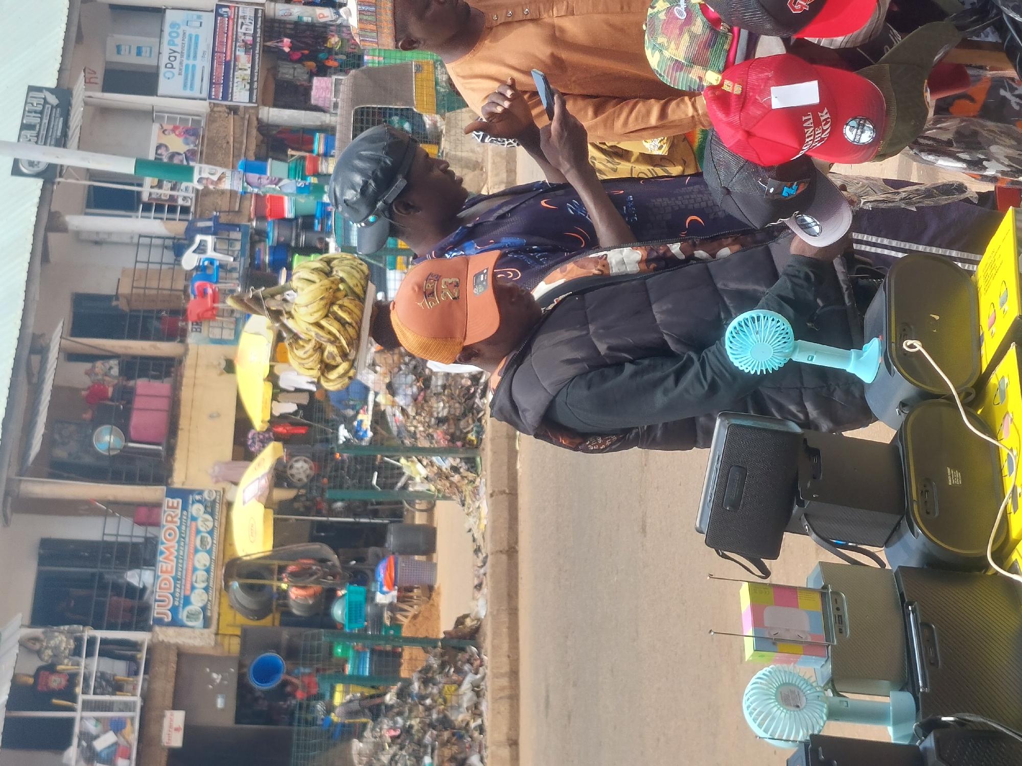In an outdoor market setting, three men are visible. The man on the left, wearing an orange baseball cap and a black puffer vest over a patterned shirt, is looking down. To his right, a man in a black baseball cap and a dark patterned shirt is holding and looking at a smartphone. The man on the far right, wearing a light brown shirt and a patterned cap, is also looking downwards, his hands are near the smartphone. In the foreground, a display of portable fans and speakers is set up. Behind the people, a street scene unfolds with various market stalls and buildings. Piles of refuse are visible on the left and right sides of the frame, indicating a less than pristine environment. In the background, a vendor is carrying a large bunch of bananas on their head. The overall scene depicts a bustling marketplace with a focus on the interaction between the three men and the merchandise.