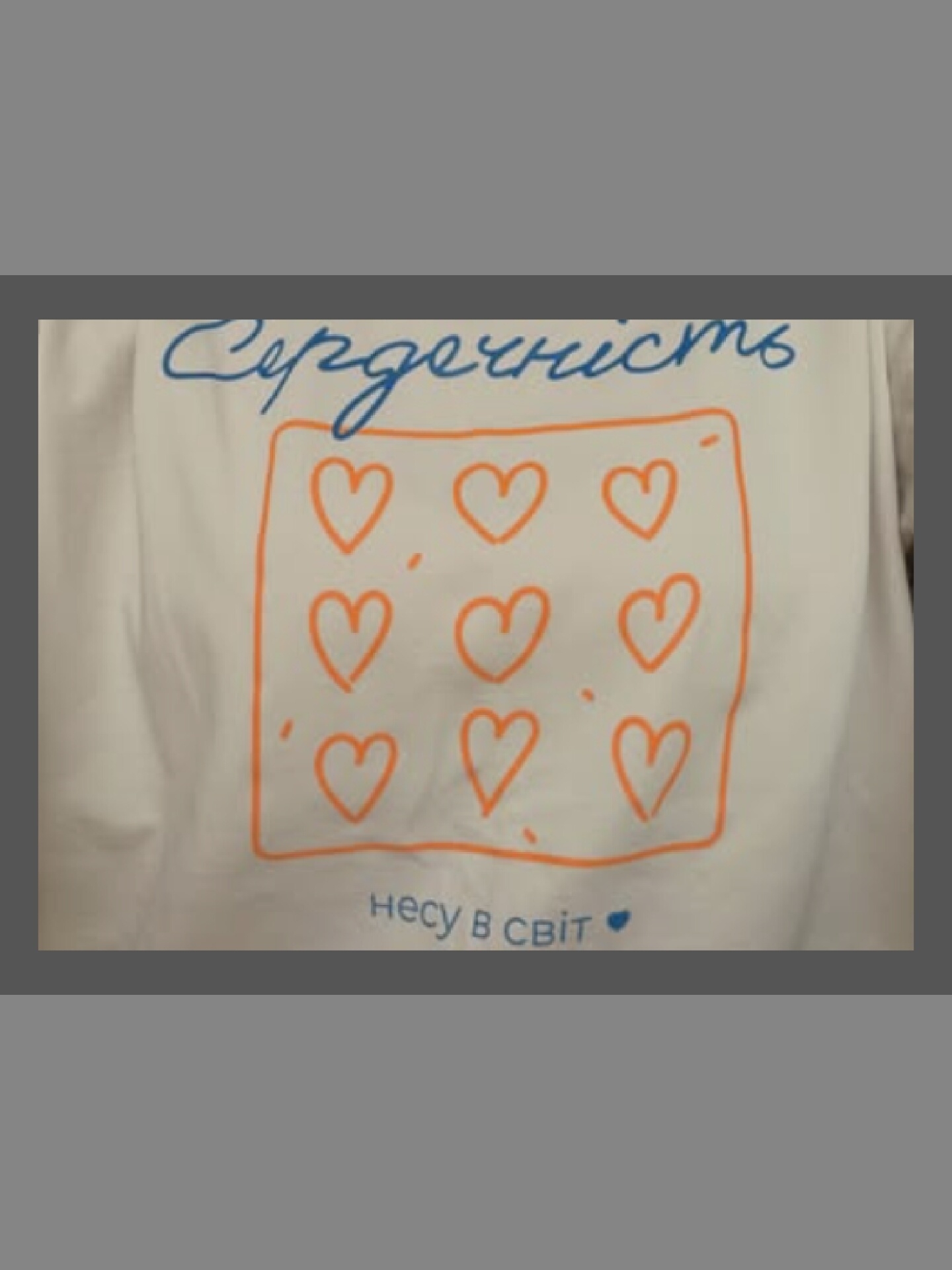 A white t-shirt displays blue cursive text at the top, followed by a grid of nine orange hearts enclosed in a square outline. Below the heart grid, there is more blue text and a small blue heart. The t-shirt is laid flat against a grey background.