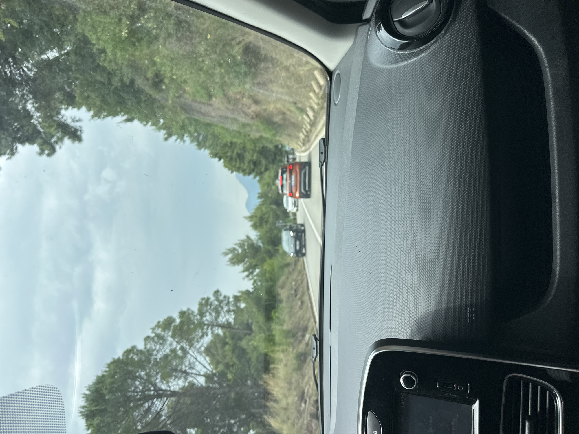 The image captures a view from inside a car, looking forward through the windshield onto a road. The car's dashboard, with its textured surface, an air vent, and part of an infotainment system, occupies the right side of the frame. Through the windshield, a line of traffic is visible, with a distinctive red SUV or crossover leading the queue, followed by a dark-colored car and others further down the road. A white concrete guardrail runs along the right edge of the road ahead.
The scene depicts a winding road passing through a lush, green landscape characterized by abundant trees and foliage on both sides. The sky is overcast with visible clouds, suggesting a cloudy but bright daytime. The setting appears to be a scenic route, consistent with the location context of Sveti Stefan, Montenegro, likely indicating a mountainous or coastal road.
The activity taking place is driving or being a passenger in a vehicle, stuck in traffic. Notable details include the time "14:26" displayed on the infotainment screen, indicating it is 2:26 PM. The text "SRS" (Supplemental Restraint System) is embossed on the dashboard.