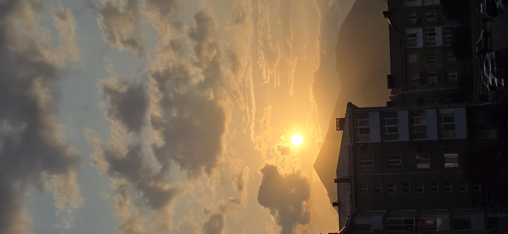 A vertical, mid-range shot captures a sunset over Inozemtsevo, Russia. The sky is dominated by dramatic clouds, tinged with orange and yellow hues from the setting sun, which appears as a bright, circular orb partially obscured by a cloud formation. In the foreground, the silhouettes of apartment buildings are visible, their dark facades contrasting with the illuminated sky. A mountain range forms the backdrop, its peaks softened by the distance and atmospheric haze. The scene is devoid of any people or discernible activity.