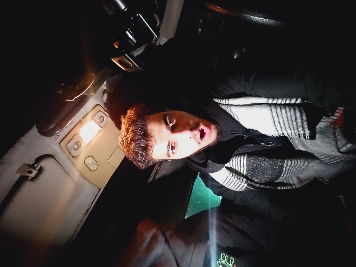 A selfie of a man inside a car with the interior light on. He has brown hair, olive skin, and brown eyes. He's wearing a black jacket and a white and grey striped scarf. The car's ceiling light illuminates his face. The dashboard and steering wheel are partially visible, with dark seats. The image has low lighting aside from the ceiling light.