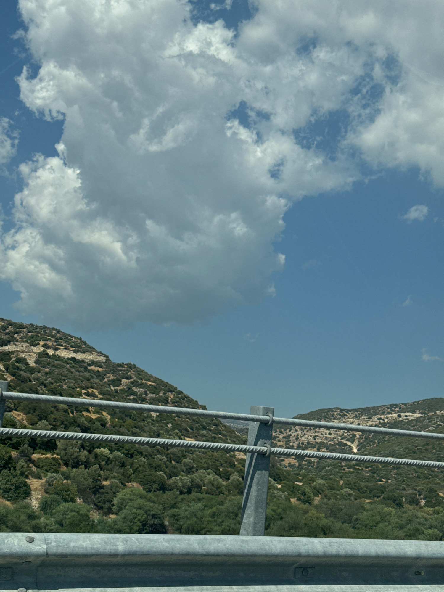 The image shows a view from a roadside guardrail, looking towards a hilly, vegetated landscape under a cloudy blue sky. The foreground is dominated by a silver metal guardrail. Behind the guardrail, the terrain rises steeply, covered with a dense mix of green trees and shrubs, with patches of exposed earth and rock. Further in the distance, the hills continue, their slopes also covered in vegetation. The sky is partly cloudy, with large, white cumulus clouds dominating the upper portion of the frame, contrasting with the clear blue sky visible in patches. The scene appears to be a natural landscape near a road.