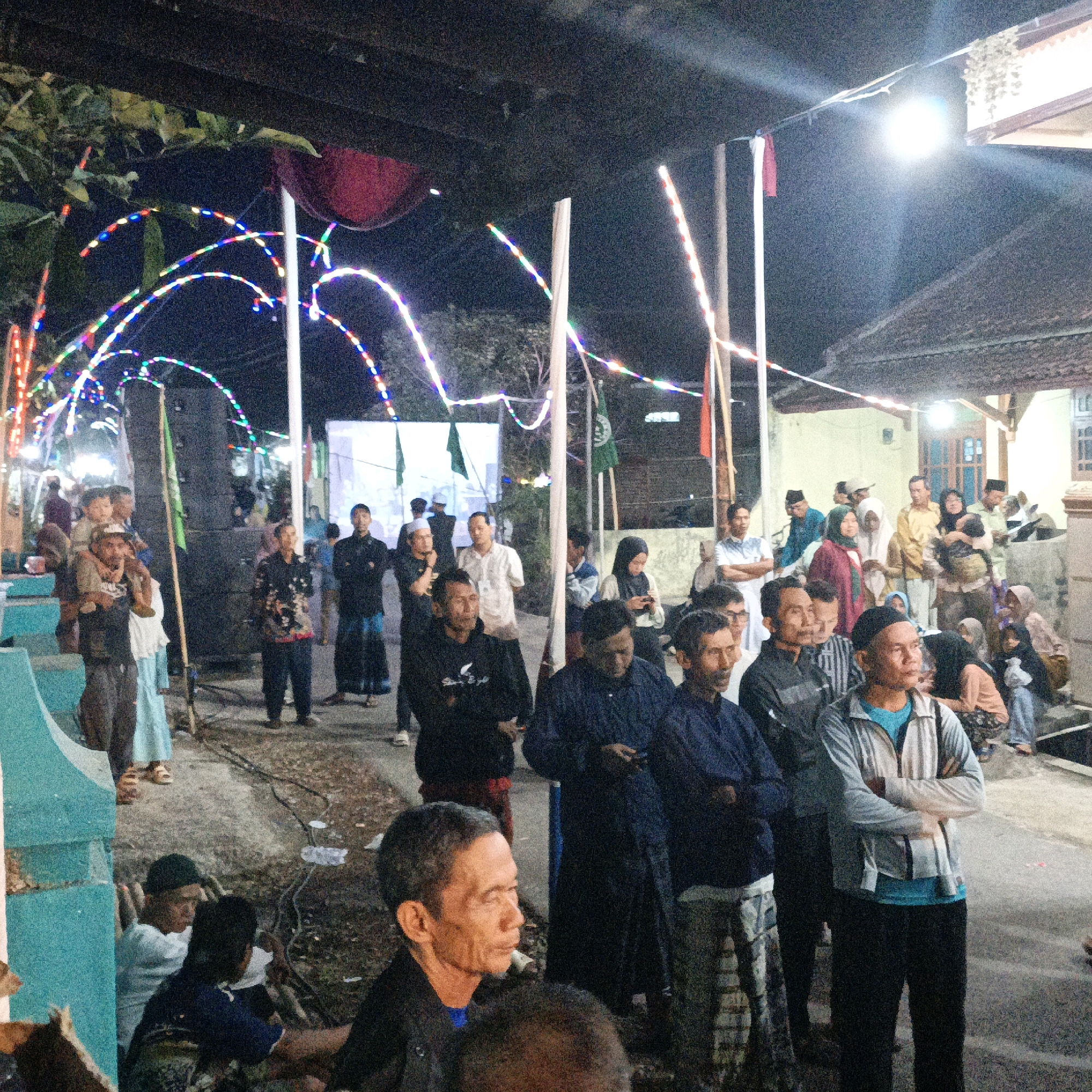 In the city of Kalitorong, Indonesia, a night gathering is depicted with illuminated string lights forming arches above a crowd of people. The scene is lit by overhead lights and the colorful string lights, creating a festive atmosphere. Several individuals are visible, some standing, others sitting or carrying children. Many are dressed in traditional Indonesian attire, including sarongs and head coverings. A large speaker is set up, suggesting a possible event or celebration. The ground appears to be dirt or gravel. The overall impression is of a community event taking place outdoors at night, possibly a religious or cultural celebration, given the attire and flags visible.