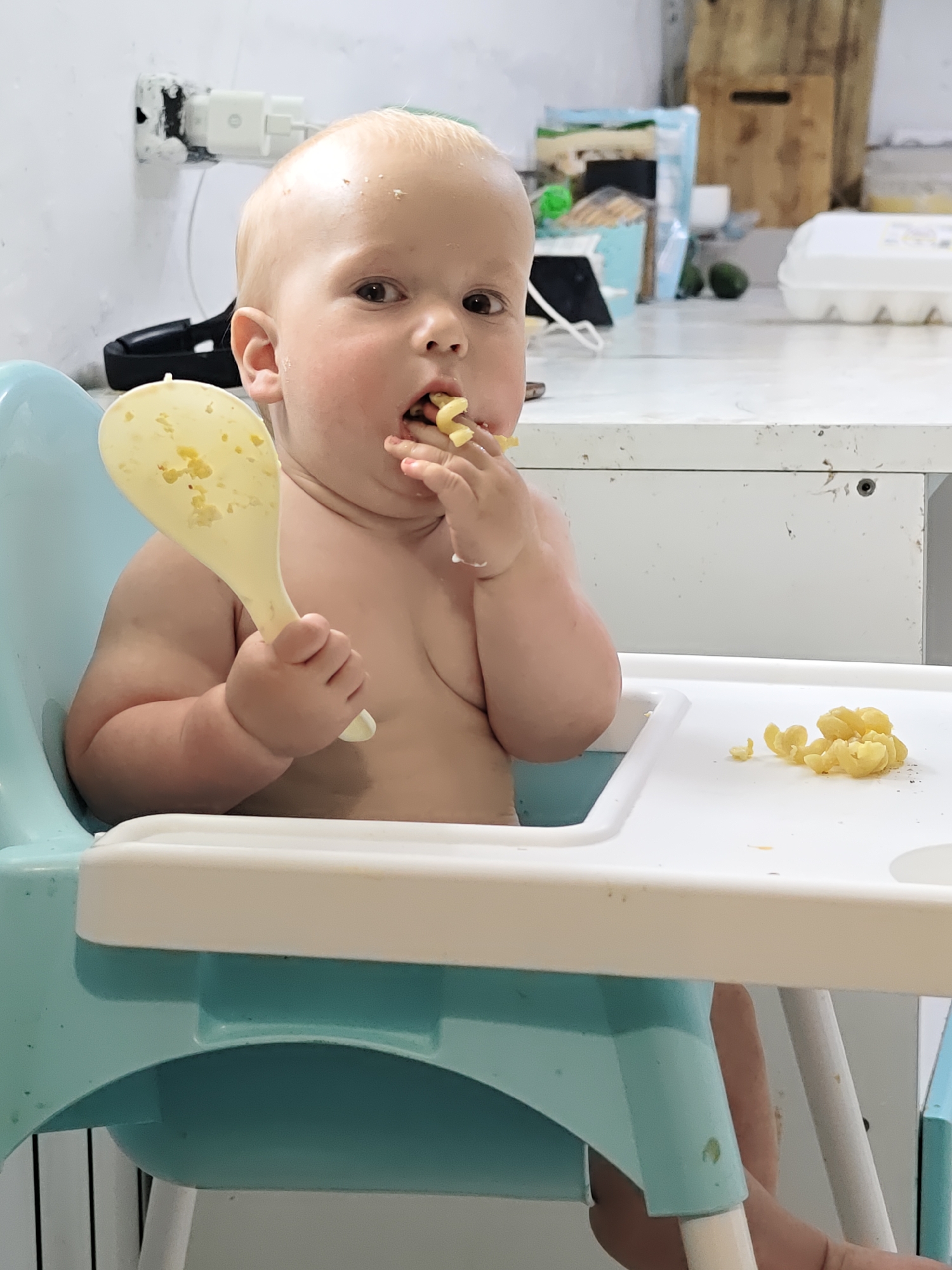 A toddler sits in a blue and white highchair, eating pasta. The toddler is fair-skinned with a light complexion and is shirtless. The toddler is holding a large yellow spoon in one hand and eating pasta with the other hand. Some pasta is scattered on the white tray of the highchair. The background includes a white wall with an electrical outlet, a countertop with various items, and a white cabinet below. The location is not Noviy, Russia.
