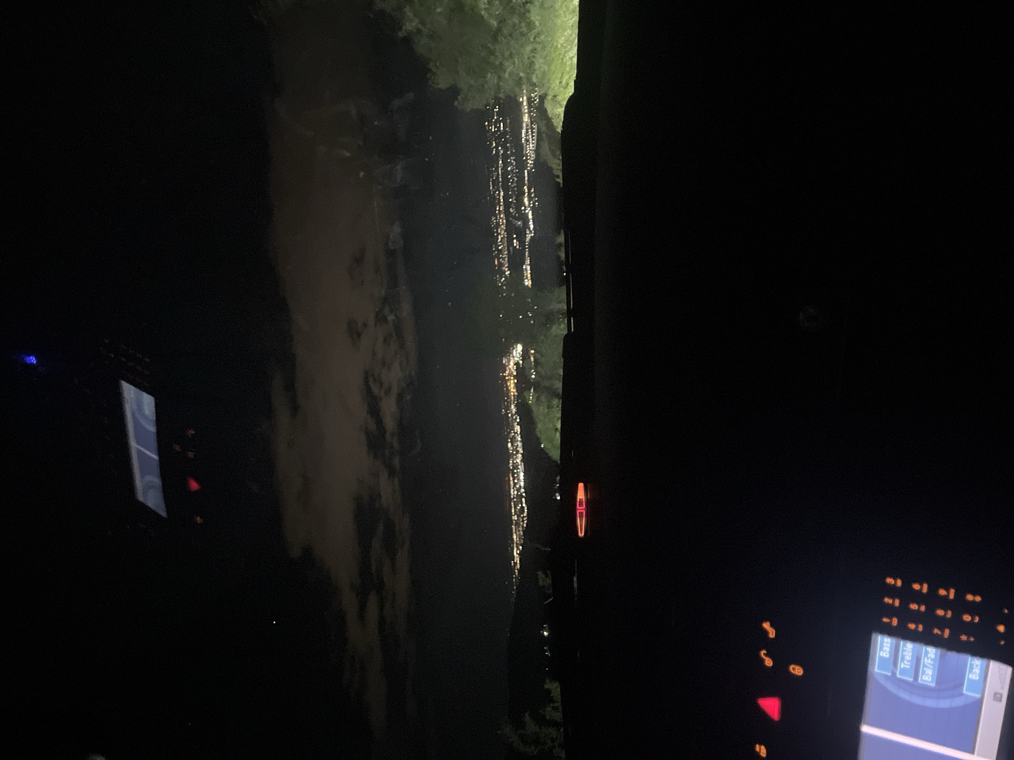 The image appears to be taken from inside a vehicle at night, showing a view through the windshield. Outside, there is a body of water reflecting the lights of a city or town. Some trees are visible along the edge of the water. The interior of the car is dark, but details of the dashboard and center console are visible, including some illuminated buttons and a screen. Based on the available information, determining the precise location within the city of Asenovgrad, Bulgaria is not possible.