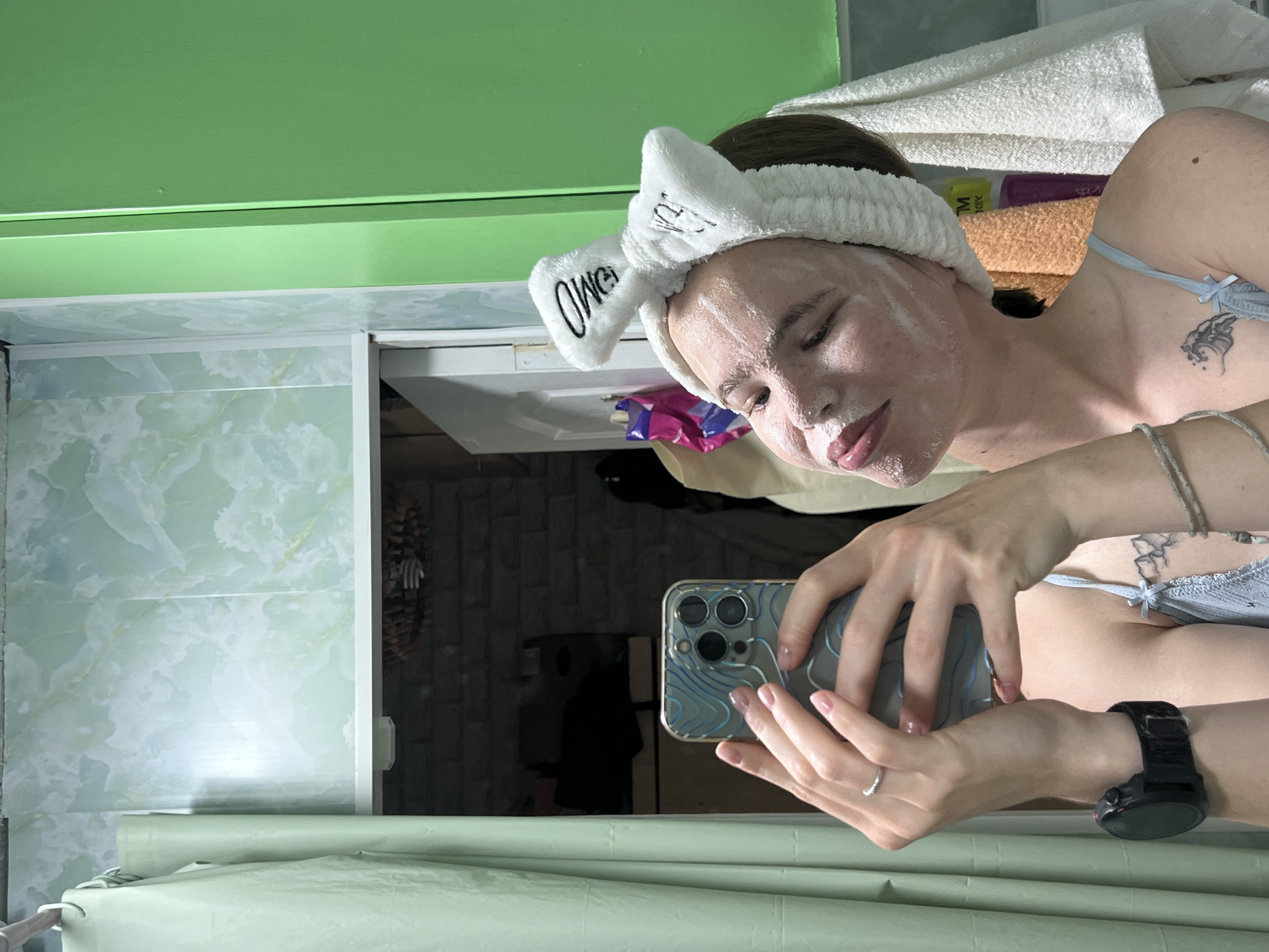 The image depicts a young woman in a bathroom. She is wearing a white fluffy headband with black lettering that appears to say "OMG" and a blue camisole. Her face is covered in a white mask, and she is holding a smartphone, taking a selfie in a mirror. The background shows a green wall, a light green shower curtain, and some towels. There are no visible elements that specifically indicate the location is in Chaikovsky, Russia.