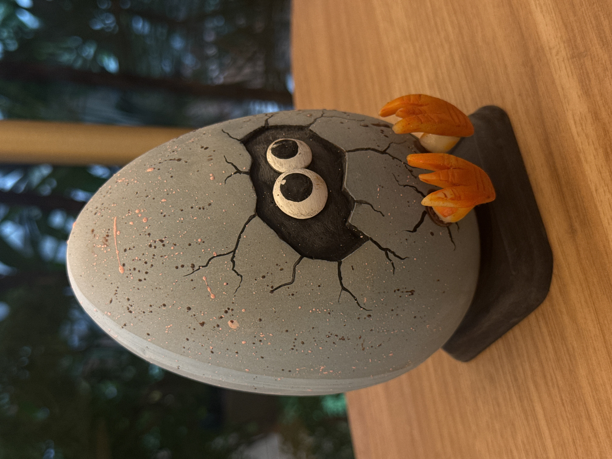 The image displays a decorative object resembling a speckled egg, horizontally oriented, with a creature seemingly hatching. The egg is primarily grey, covered in small dark brown and pinkish-red speckles, and features numerous painted black cracks across its surface. A central, irregular opening towards the left reveals a dark interior from which two distinct, white, oval eyes with large black pupils emerge, stacked vertically. These eyes give the creature a cartoonish and curious expression. To the right side of the egg, two bright orange, three-toed feet, akin to those of a bird or dinosaur, protrude from the cracked shell.
The hatching egg decoration rests on a dark, flat base, which in turn is placed against a light brown wooden surface, possibly a wall or furniture, showing visible grain. The background is softly blurred, revealing dark green foliage and trees, along with out-of-focus lighter elements and a horizontal yellow bar. This suggests an outdoor or semi-outdoor setting visible from the location, which is North Kuta, Indonesia. The object depicts the whimsical activity of a creature breaking free from its egg. The lighting indicates it is daytime, possibly indoors or in a shaded area. There is no visible text in the image.