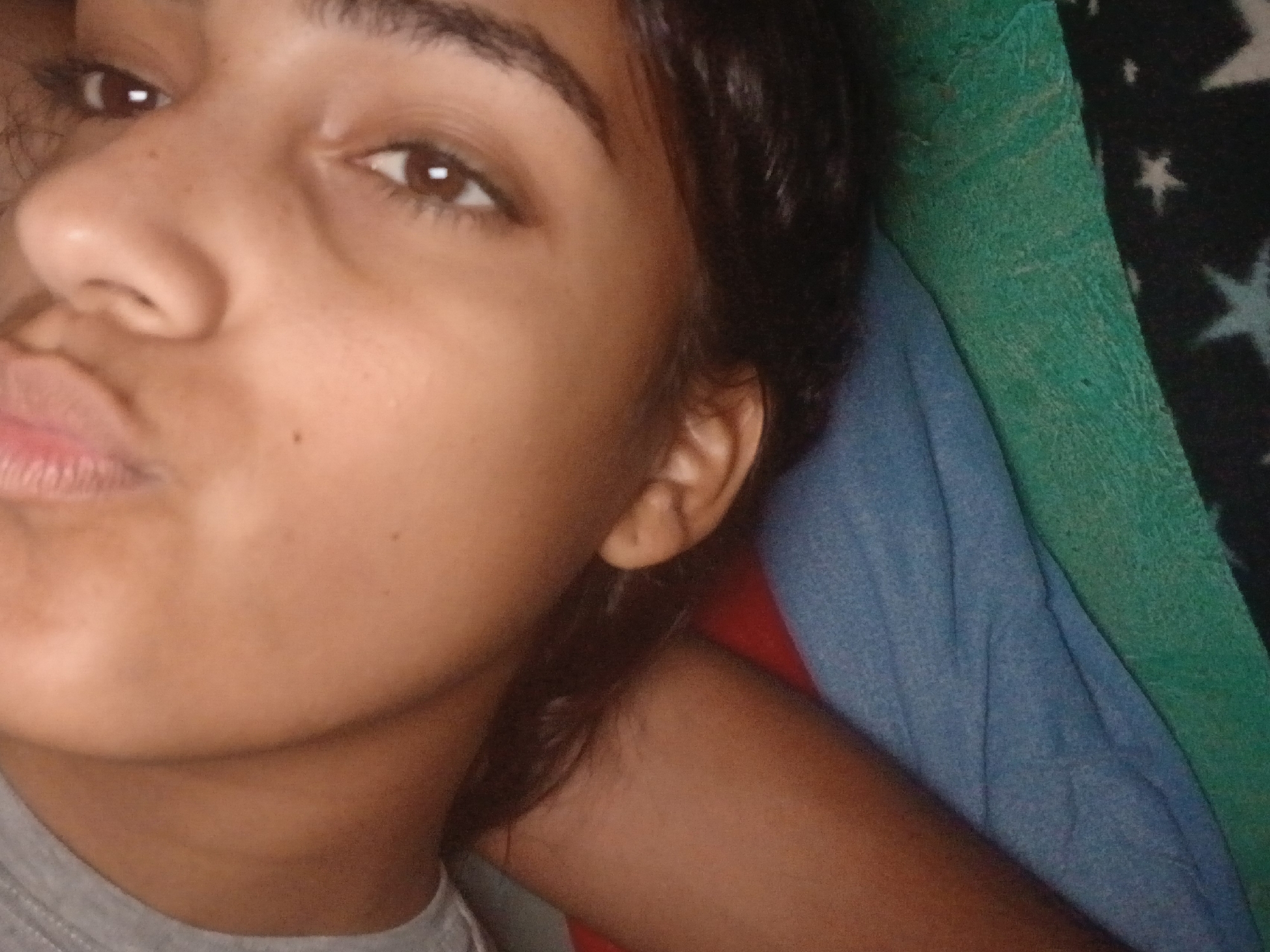 A close-up selfie of a young woman with brown eyes and dark hair. She has brown skin and is wearing a grey top. Her lips are slightly pursed. She is lying on a surface covered with blue, green, and black fabrics that have a star pattern. The location is not discernible.