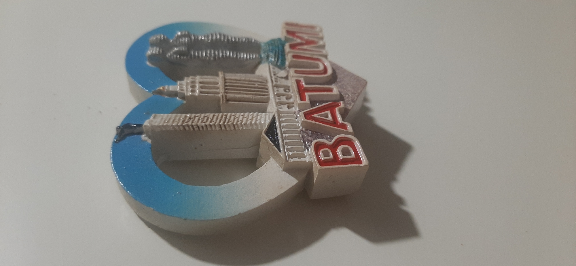 A souvenir magnet is attached to a light-colored, possibly white, vertical surface. The magnet is heart-shaped and features a relief of several buildings, resembling a city skyline. The buildings are primarily light gray or beige with some darker detailing. In the foreground, the word "BATUMI" is prominently displayed in red lettering with white outlines. The background of the magnet is a gradient of blue and white, suggesting a sky. A shadow is cast by the magnet onto the surface it's attached to, indicating the presence of a light source from the left and slightly above. The image is taken from a slightly angled perspective, with the top left of the magnet closer to the viewer. There is no indication of Yessentukskaya, Russia in the image; the text clearly reads "BATUMI," which is a city in Georgia.