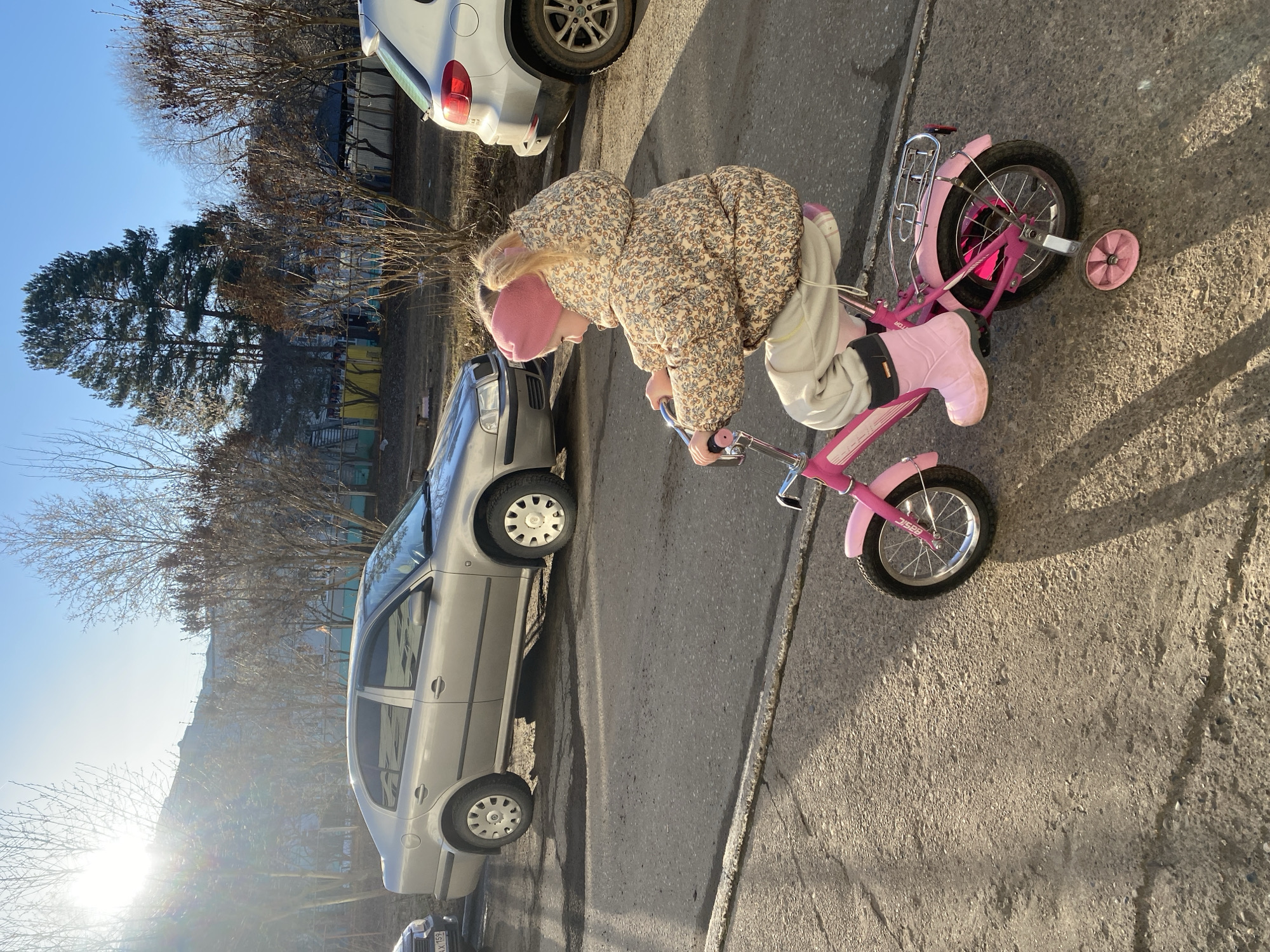 A young child, presumably a girl, is on a pink bicycle with training wheels on an asphalt road. She is wearing a floral-patterned jacket, light-colored pants, pink boots, and a pink hat. A silver car is parked on the left side of the road. A white car is also visible parked nearby. In the background, there are trees and buildings. The location is the city of Chaikovsky, Russia, based on the license plate. The lighting indicates it is daytime, with some sunlight casting shadows.