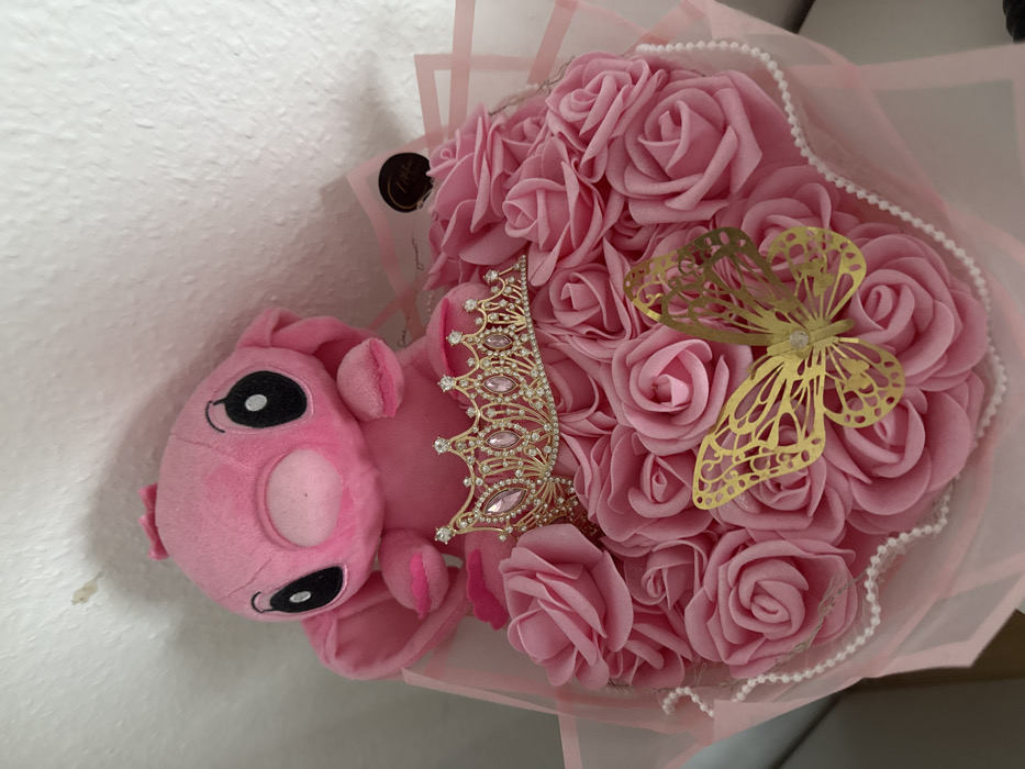 A gift bouquet is displayed in an indoor setting in Vellmar, Germany, positioned against an off-white textured wall exhibiting minor discoloration in the upper left. The central component is a large cluster of uniformly sized pink artificial roses. A pink plush toy, resembling the character Angel, is seated upright atop the rose arrangement, featuring large black eyes and a lighter pink facial area. Embedded among the roses, directly below the plush toy, is a gold-toned tiara adorned with numerous clear and pink rhinestones. A gold-colored, intricately cut butterfly ornament rests on the roses in the lower right quadrant of the bouquet. White, spherical beads are strung along the visible perimeter of the rose cluster. The entire assembly is encased in translucent pink wrapping material, which includes a small, dark circular sticker on its upper right side. No individuals are present in the frame.