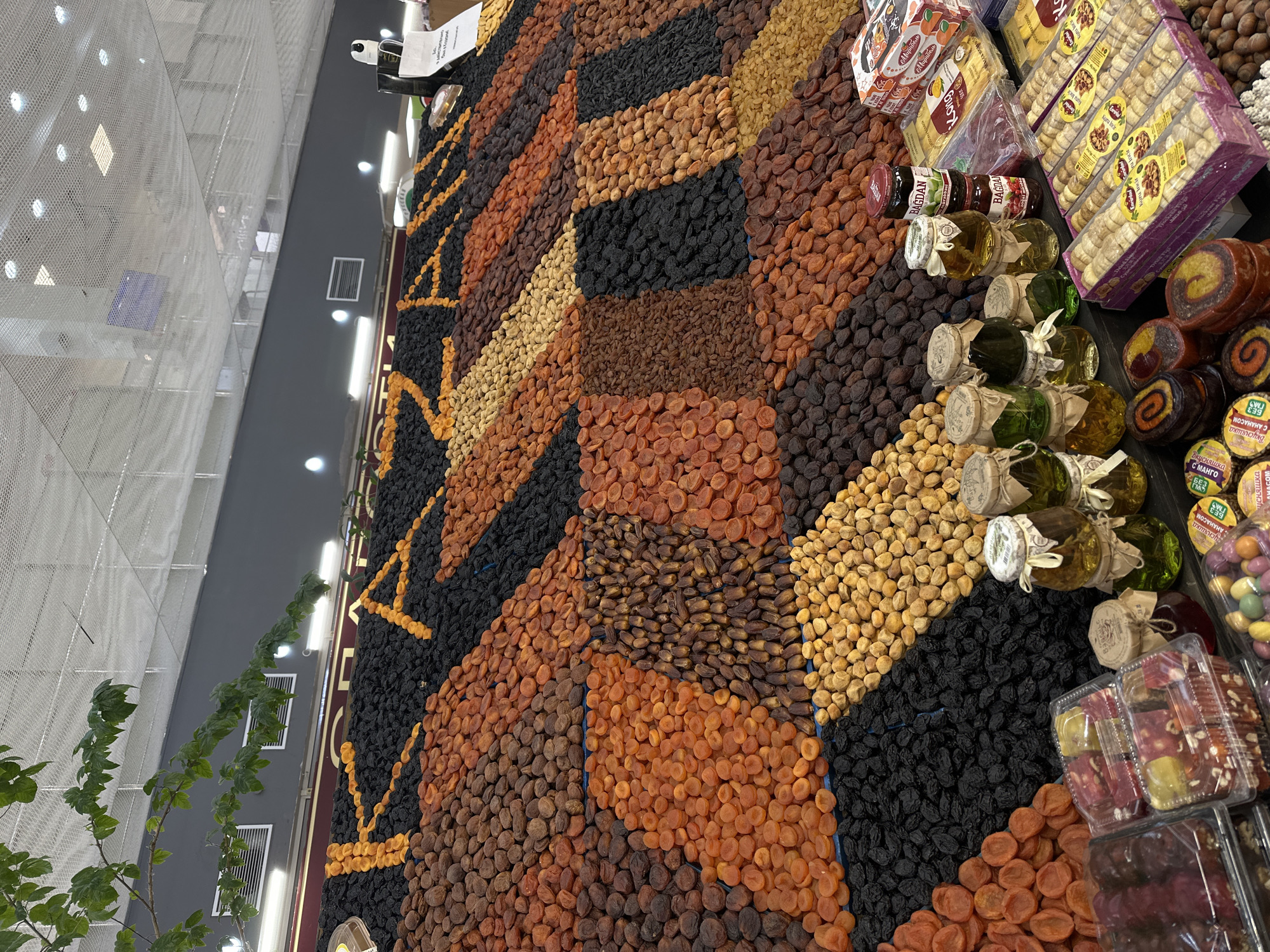 The image shows a display of dried fruits and other confectionery items in a market, likely in Kazan, Russia. The dried fruits, primarily apricots and prunes, are arranged in colorful patterns. The products for sale include dried fruits like apricots and prunes, as well as jars of what appears to be honey or preserves, and packaged sweets such as Turkish delight. The scene is lit by overhead lighting in a commercial setting. The store is near a gray wall and has a tall plant or tree. The image is taken from above, providing a view of the arrangement of goods on a table.