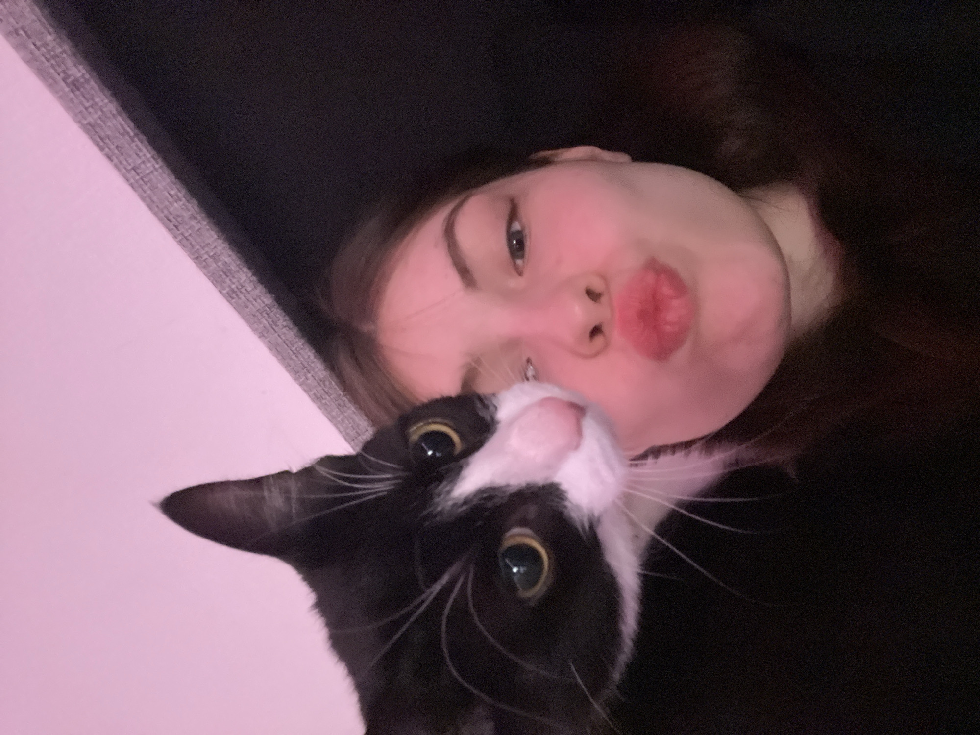 A selfie captures a young woman with long, dark hair in a dark setting, juxtaposed with a tuxedo cat. The woman has fair skin and her lips are puckered. The cat is black and white with big, dilated eyes. The background suggests an indoor setting, with a gray textured object visible. The image is taken close-up, focusing on the faces of both the woman and the cat. There is no indication of the location being Saint Petersburg, Russia in the image.