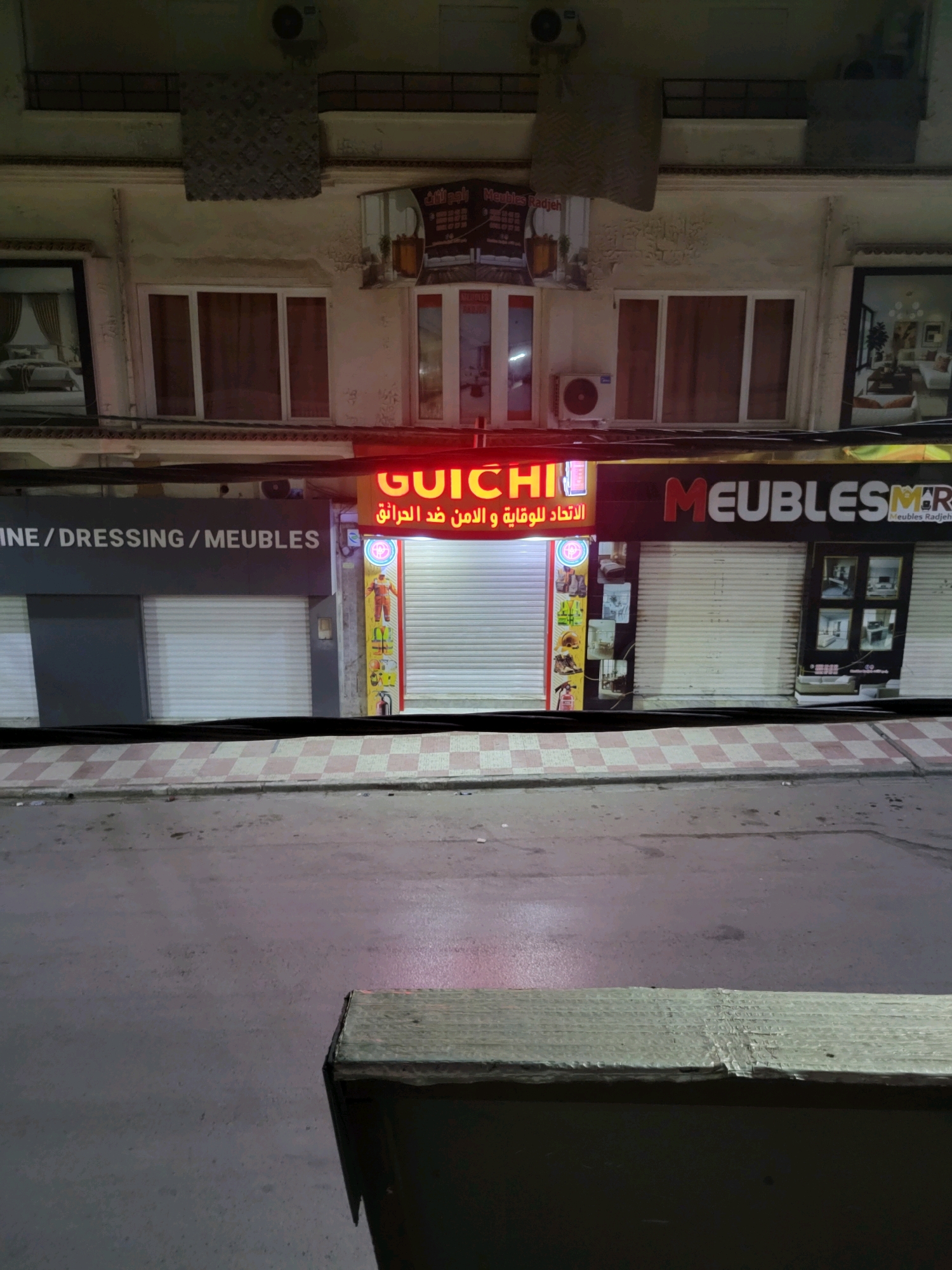 The image shows the facade of a building in the city of Bordj Bou Arreridj, Algeria, at street level. The building has multiple storefronts. One storefront, on the left, has a dark gray facade with white lettering that reads "INE / DRESSING / MEUBLES". Another storefront, to the right, has a bright red neon sign that reads "GUICHI" above Arabic text. Further to the right, there is a black sign with white lettering that reads "MEUBLESMR". The storefronts have their metal roll-up doors closed. There are images displayed on the rightmost storefront, showing furniture. The street in front of the building is wet and reflects the lights from the storefronts. A portion of a wooden structure is visible in the foreground. The overall lighting suggests it is nighttime or dusk.