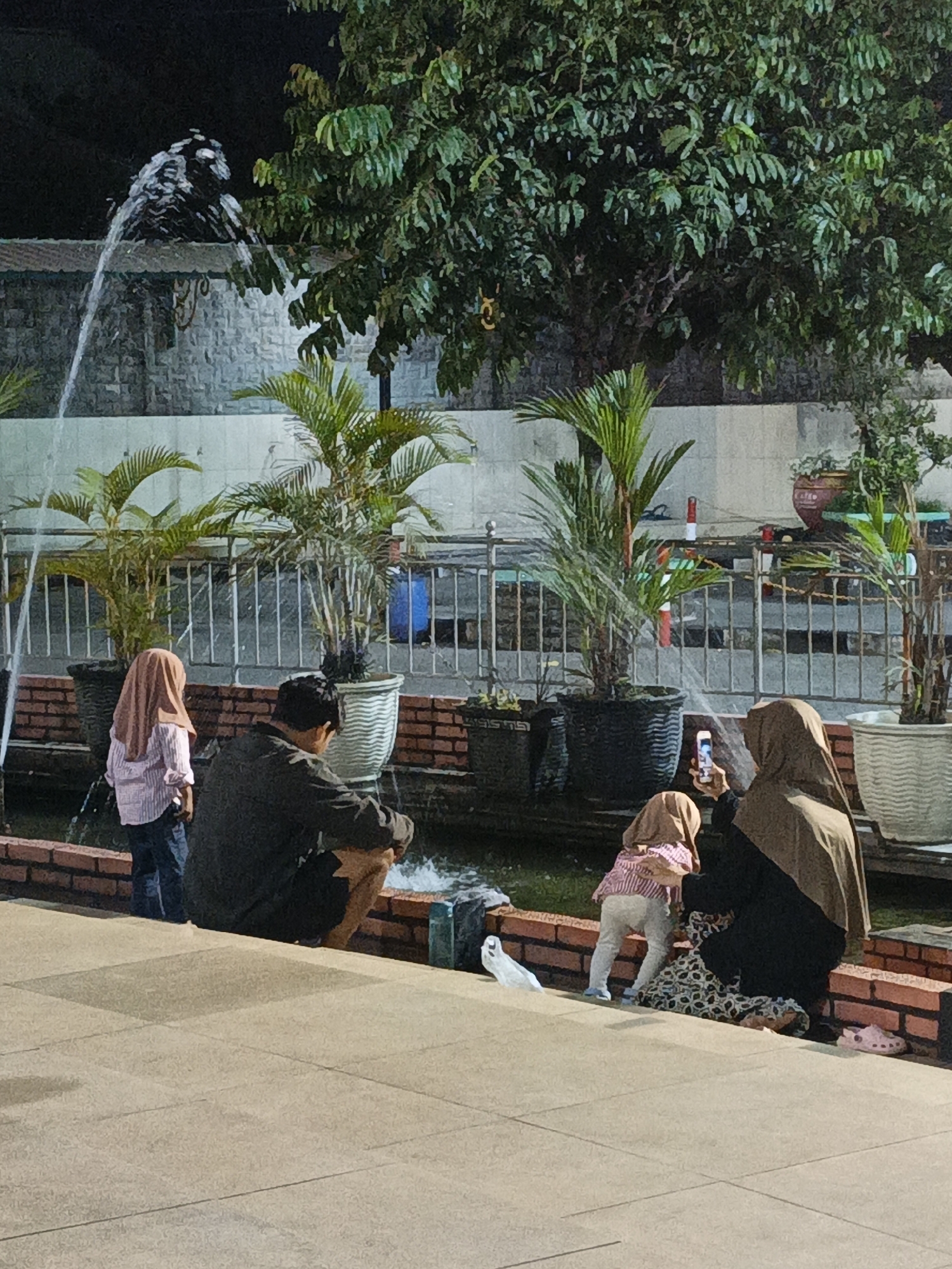 The image captures a nighttime scene at a fountain area in the city of Jonggrangan, Indonesia. In the foreground, two children, one standing and one squatting, are visible. The standing child, on the left, wears a light-colored headscarf and a striped shirt. The squatting child, on the right, also wears a headscarf and patterned pants. An adult male, wearing a dark jacket, is partially visible, squatting near the fountain. On the right side of the frame, another adult, wearing a dark headscarf and patterned skirt, is holding a mobile phone, seemingly taking a picture or video of the children.
The fountain itself is a prominent feature, with water spraying upwards and cascading into a shallow pool. Surrounding the pool are several potted plants, including palm trees, and decorative brickwork. In the background, a large tree with dense foliage is illuminated, and a wall with a textured finish can be seen. The overall lighting suggests it is evening or night, with artificial lights illuminating the fountain area. The ground in the foreground is paved with large, light-colored tiles. A pair of pink clogs are visible on the right side of the paved area.