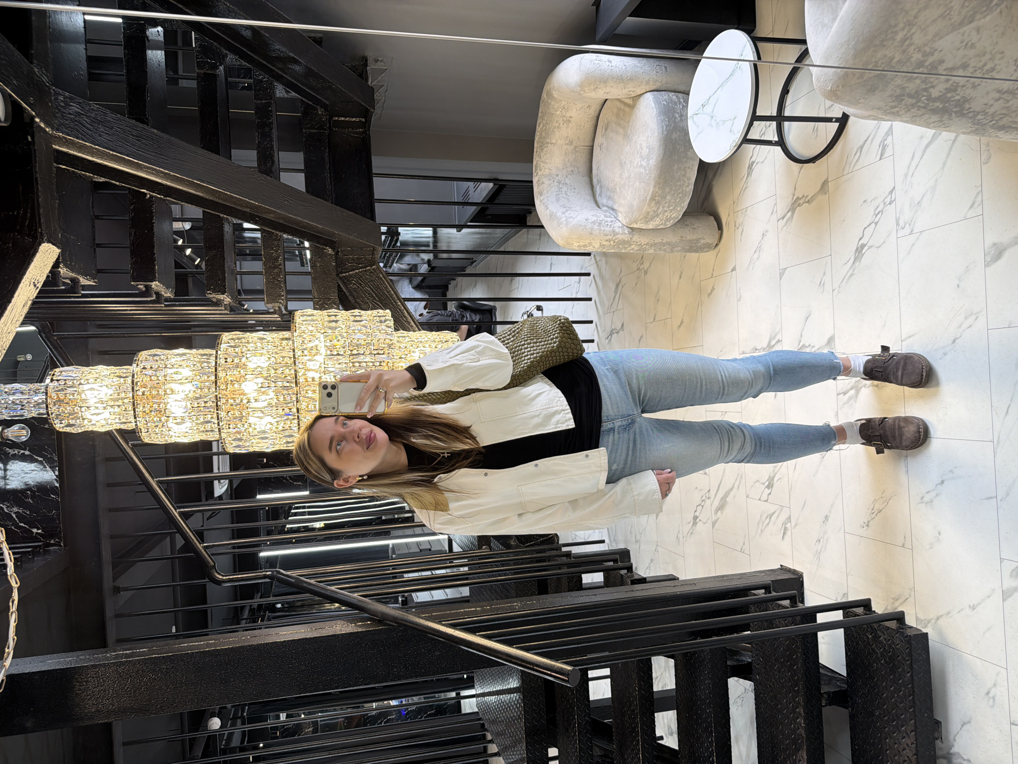 The image captures a selfie taken by a young woman in an upscale interior setting, likely in Moscow, Russia. The woman, appearing to be in her 20s or early 30s with long, light brown hair, is looking up and to her right with a playful expression, her tongue slightly extended. She is dressed in a white jacket over a black top, light blue jeans, and dark brown shoes paired with white socks. She holds a light-colored smartphone in her left hand, which has a ring on the ring finger, suggesting she is taking the photo. A textured, possibly olive green or khaki, shoulder bag rests on her right shoulder.
The background features a striking blend of industrial and luxurious elements. A grand, multi-tiered crystal chandelier, brightly illuminated, hangs prominently in the upper left, providing warm light. To the left, a dark, industrial-style metal staircase with diamond-plate steps and black railings ascends upwards. On the right, two plush, light-colored (possibly white or light grey) armchairs with a distinctive, wavy design are visible. Between them, a small, round table with a white marble-patterned top and a black metal frame adds to the modern aesthetic. The floor is covered in large, rectangular light-colored tiles with grey marble-like veining. Some dark, possibly black marble-patterned wall panels are also visible near the staircase. The overall setting suggests a contemporary lobby or lounge area within a commercial building, such as a hotel or a high-end office. There is no visible text in the image.