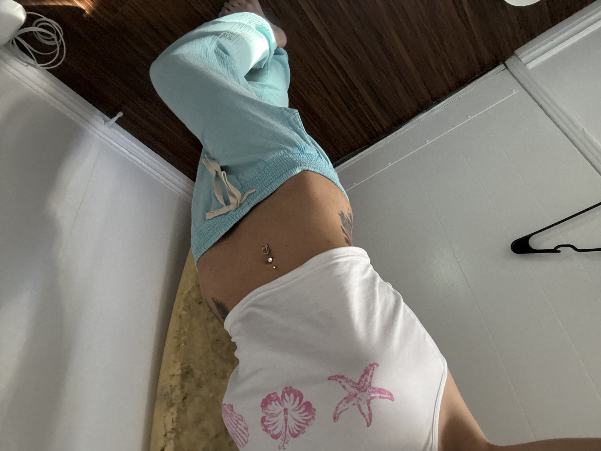 The image shows a person indoors, lying on what appears to be a cushioned surface in a room with brown wooden flooring and white walls. The person is wearing light blue pants and a white top with pink hibiscus and starfish designs. The view is from above, focusing on the person's midsection, where a belly button piercing is visible. Part of a clothes hanger is seen on the white wall. The location and any activities are not clear from the image. There is no definitive information about Seal Beach, United States of America in the image.
