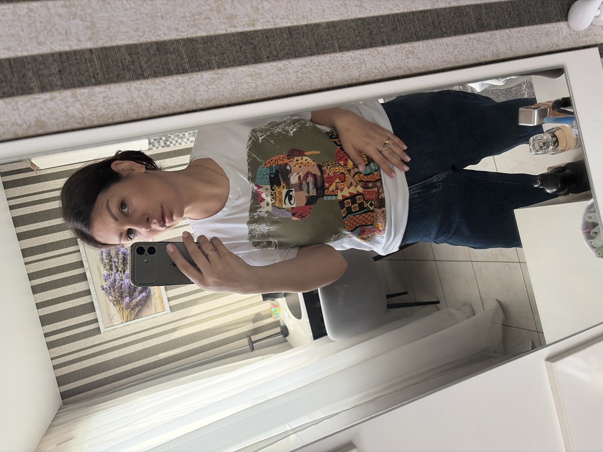 A female, identified as residing in the city of Sochi, Russia, is captured in a mirror selfie. She has dark hair pulled back, a light complexion, and is wearing a white short-sleeved t-shirt featuring a multi-colored abstract portrait graphic. She is also wearing dark blue jeans. Her right hand holds a dark gray smartphone with two visible camera lenses, angled to reflect her image in the mirror. Her left hand rests on her lower abdomen, displaying a gold-toned ring on her ring finger and light pink manicured nails.
The background, visible through the mirror's reflection, reveals an indoor setting. To the left, a window with sheer white curtains allows ambient light. The wall behind the woman features light beige vertical stripes against a darker background. A framed picture depicting a bouquet of purple lavender is mounted on this wall. Further to the left, a portion of a room includes a black desk with a circular top, a gray upholstered chair, and a black desk lamp. A clear bowl containing small, colorful objects is also visible on the desk. The floor is covered with light-colored square tiles. Below the mirror, on a surface, several items are partially visible, including perfume bottles and a dark figurine.