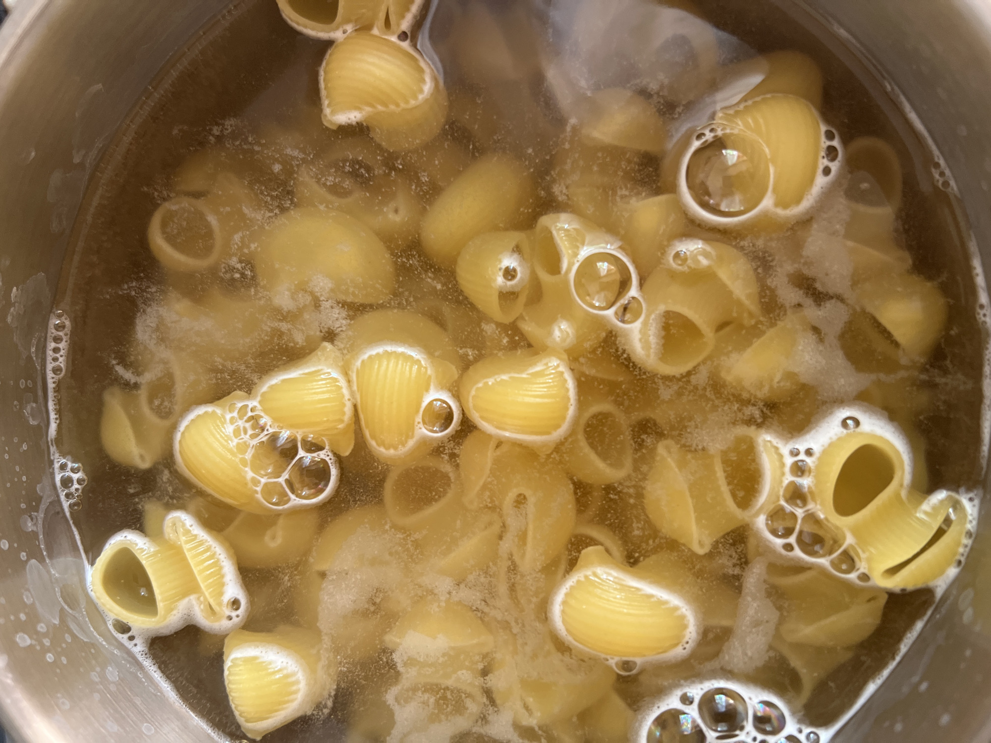A close-up, overhead shot shows conchiglie pasta being boiled in a metal pot. The pasta is yellow and has a ridged texture, with many pieces submerged in cloudy, bubbling water. Some pieces are floating, partially submerged, and covered in foam. The pot appears to be stainless steel, with a slight sheen reflecting the light. The lighting suggests an indoor environment, possibly a kitchen. There are no people or other objects visible in the frame.