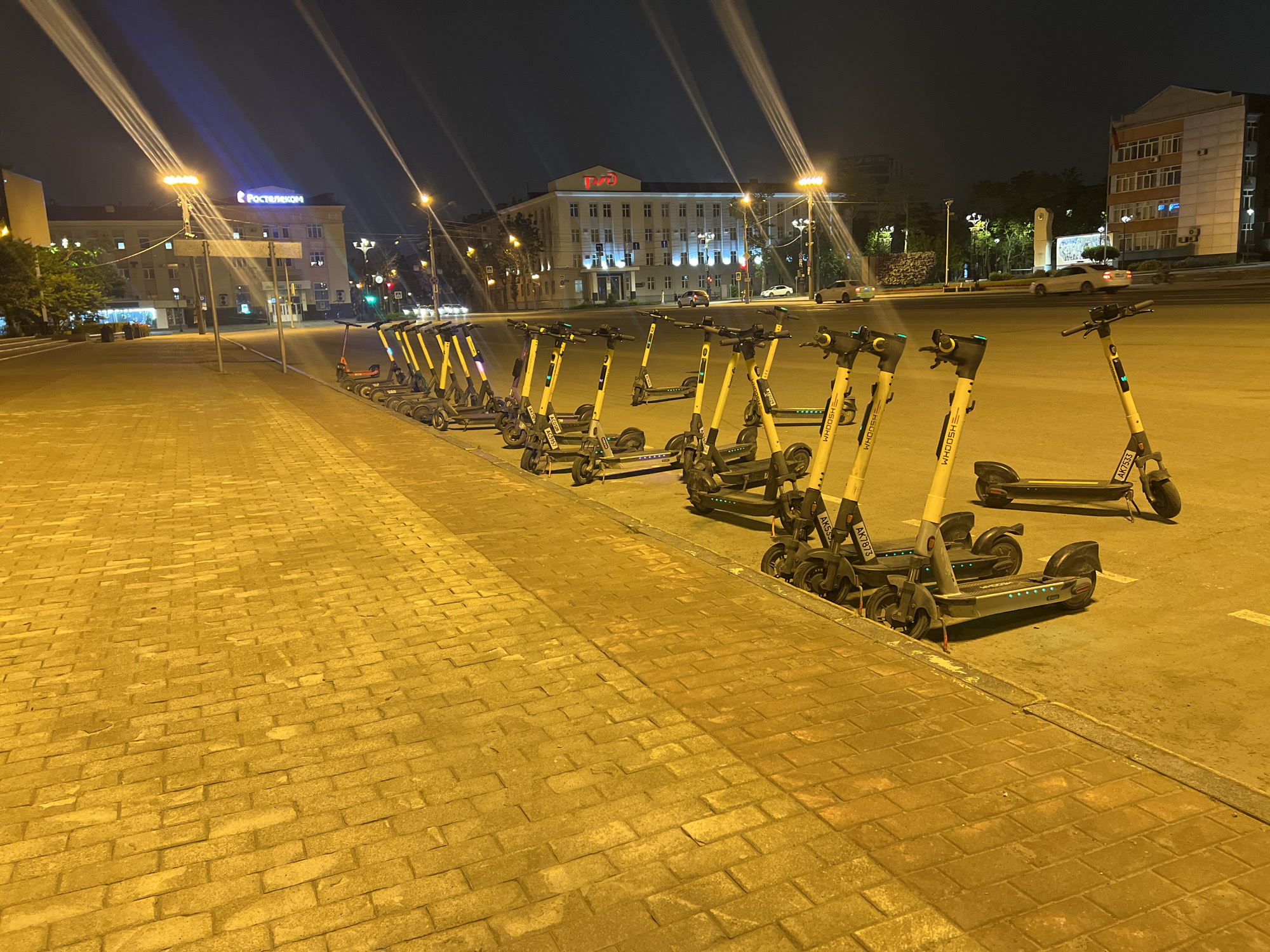 The image captures a street scene at night in Yuzhno-Sakhalinsk, Russia. A row of electric scooters is parked on a paved area, illuminated by streetlights. The scooters are predominantly yellow and gray. In the background, buildings are visible, including one with a prominent sign that reads "Ростелеком" (Rostelecom) in blue letters. Another building displays a red sign that appears to be "РЖД" (RZD - Russian Railways). Vehicles are present on the road in the distance. The ground in the foreground is paved with yellow bricks. The overall lighting suggests an urban environment at night, with artificial illumination casting long shadows.