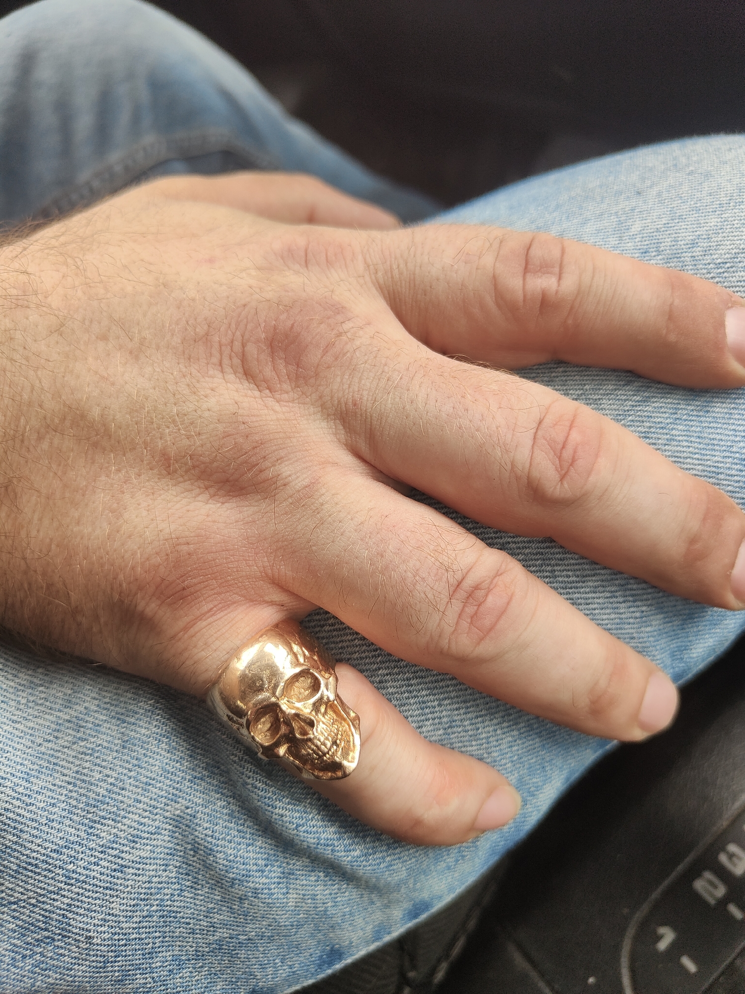 The media file shows a close-up of a person's hand resting on their lap, which is covered in blue denim jeans. The hand is adorned with a gold-colored ring shaped like a skull, worn on the index finger. The ring is detailed, with visible eye sockets, a nose cavity, and teeth. The skin on the hand appears fair and has visible pores and fine hairs. The knuckles and finger creases are distinct. The lighting suggests it may be daytime, and the background is blurred, obscuring any specific location details. The overall mood is neutral, with no apparent emotional cues conveyed by the hand or the ring. No text is visible in the image.
